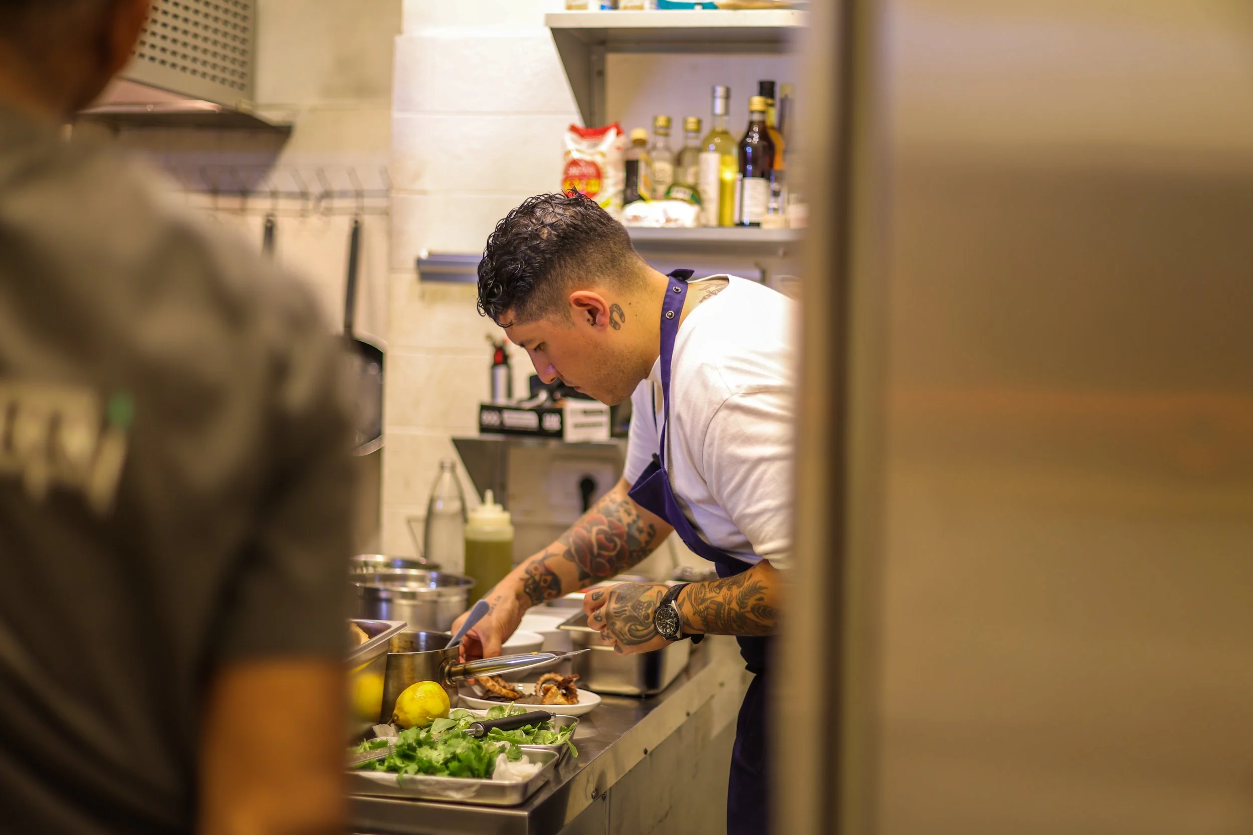 A tattooed male chef preparing ingredients in a professional kitchen. He is wearing a white shirt, a purple apron, and watches. The counter has fresh greens, lemons, and kitchen utensils.
