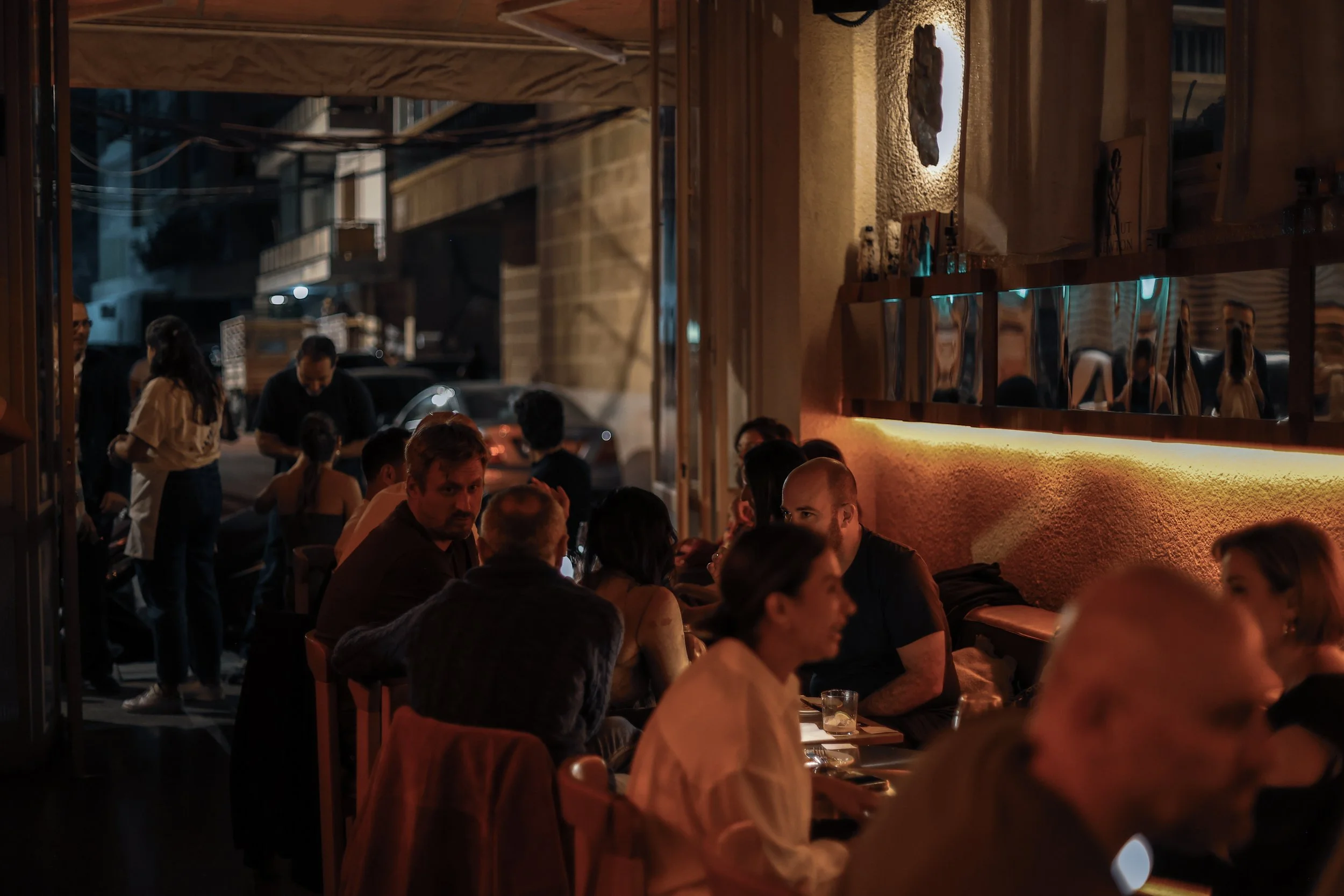 People dining inside a restaurant at night, with some patrons sitting at tables and others standing near the entrance, warmly lit with orange lighting and street view outside.