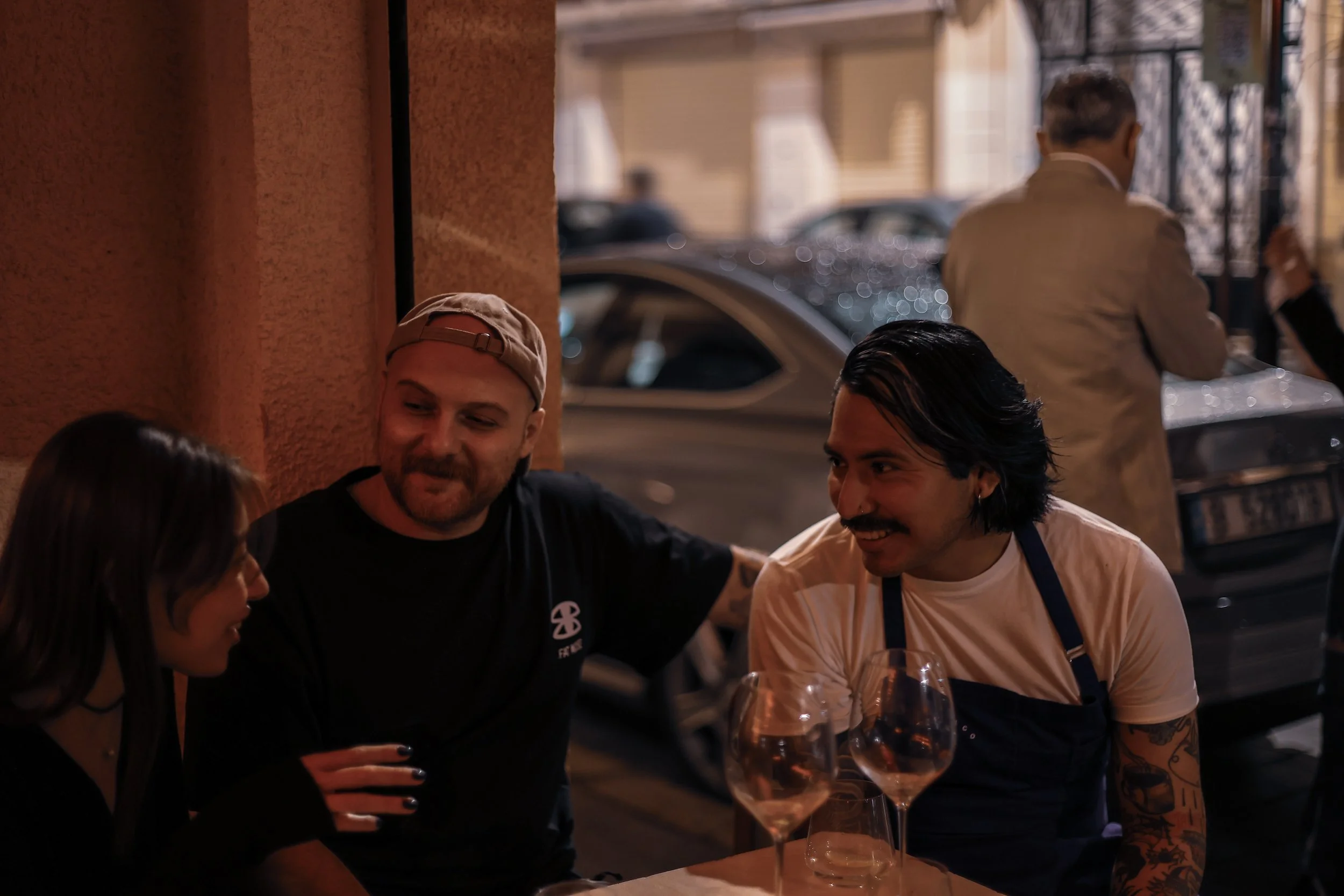 Three people sit at an outdoor table enjoying drinks at night, engaging in conversation, with cars and other people visible in the background.