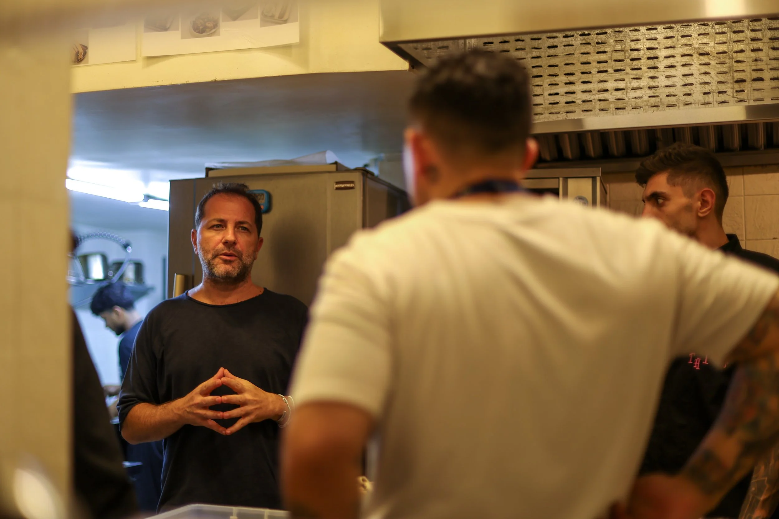 A man with dark hair and a beard, wearing a black shirt, is speaking to three other men in a restaurant kitchen. Four of the men are visible, with the man facing the camera and gesturing with his hands, while the other three are facing away or to the
