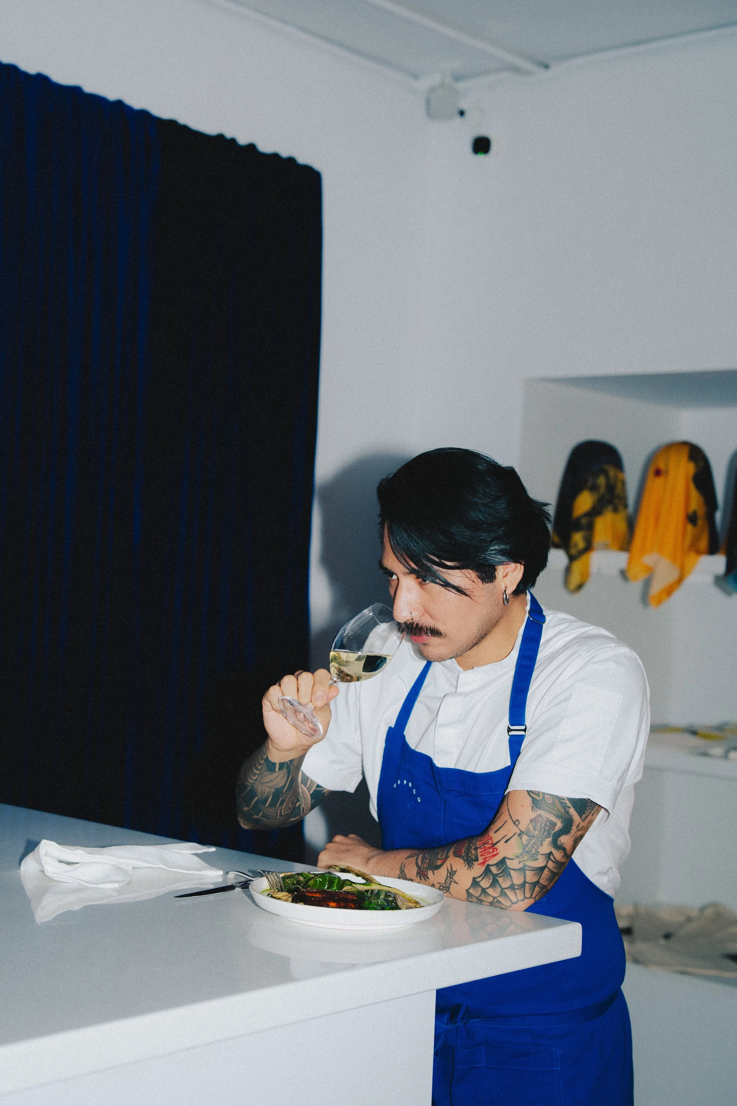 A chef with tattoos tasting white wine in a modern kitchen.