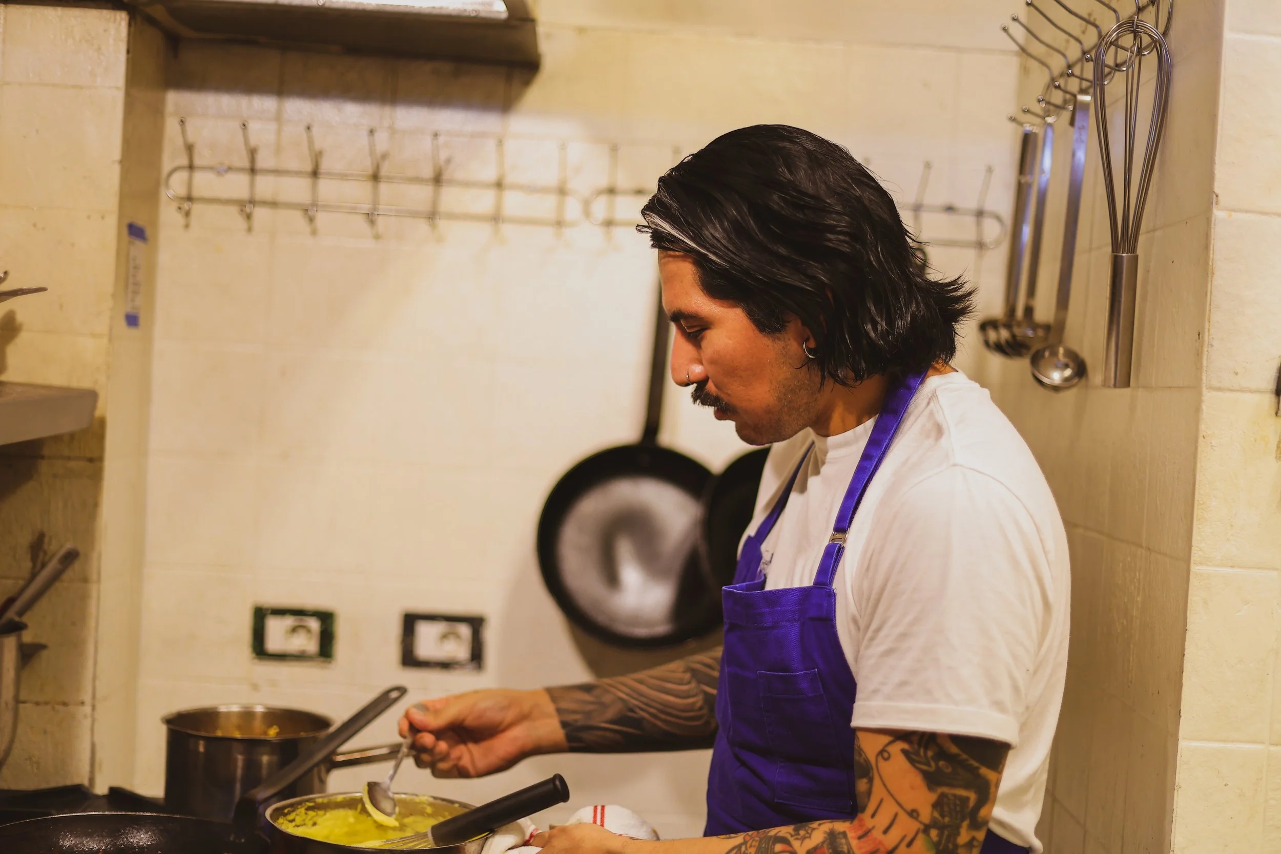 A man with black hair, tattoos on his arms, and wearing a white shirt and blue apron cooking in a kitchen.