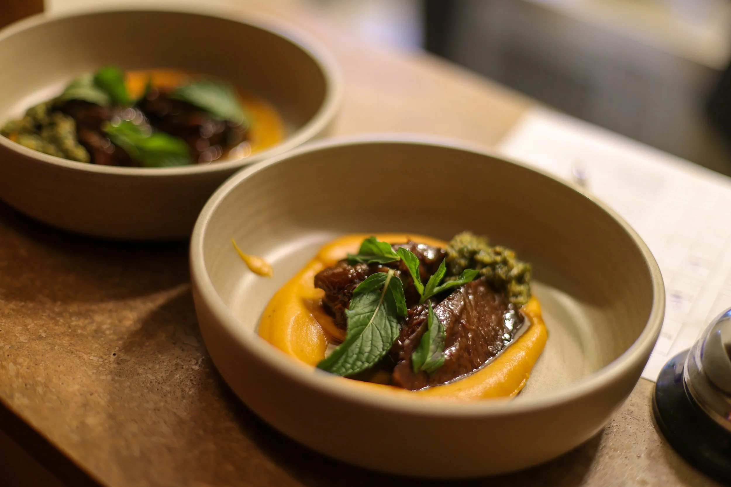 Close-up of two beige bowls filled with a gourmet beef dish garnished with herbs, resting on a wooden surface.