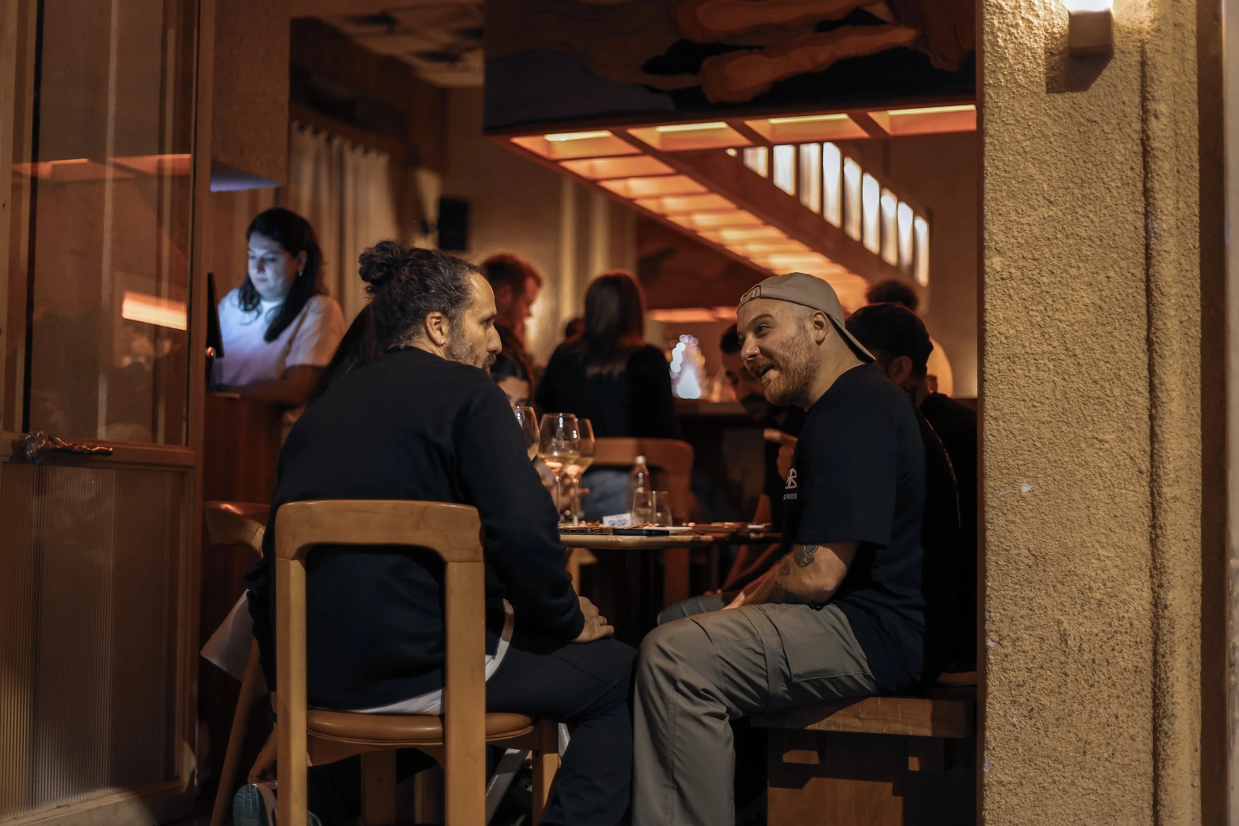 Two men sit at a table in a dimly lit restaurant, engaged in conversation. One man has long dark hair tied back, and the other wears a backwards baseball cap. There are glasses of wine on the table, and other diners are visible in the background.