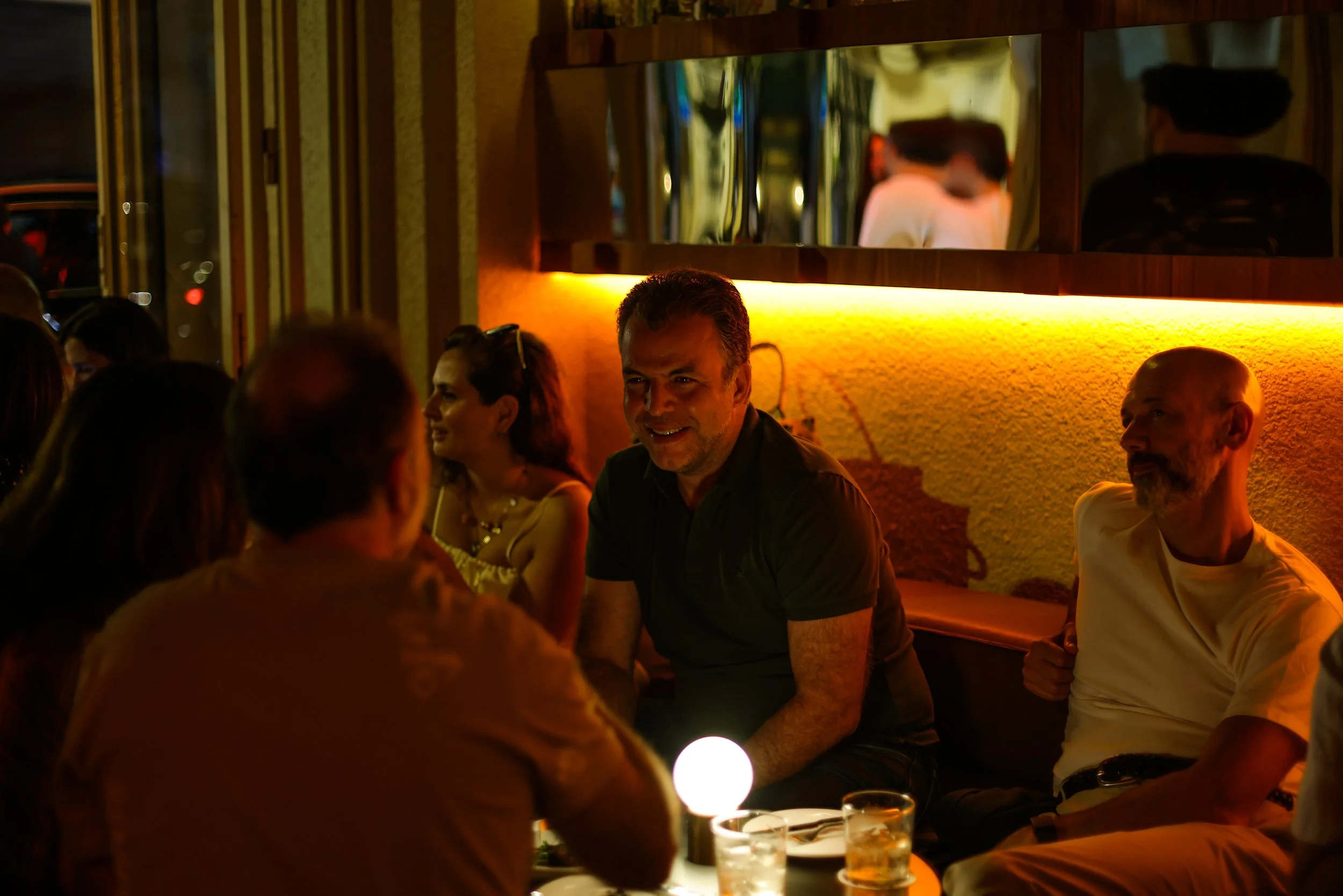 Group of people dining in a dimly lit restaurant, smiling and talking, with colorful lighting and modern decor.