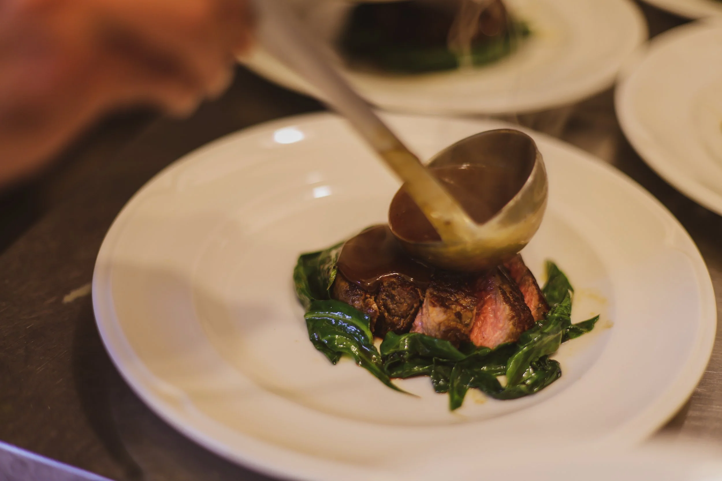 A chef pouring sauce over a cooked steak topped with greens on a white plate.