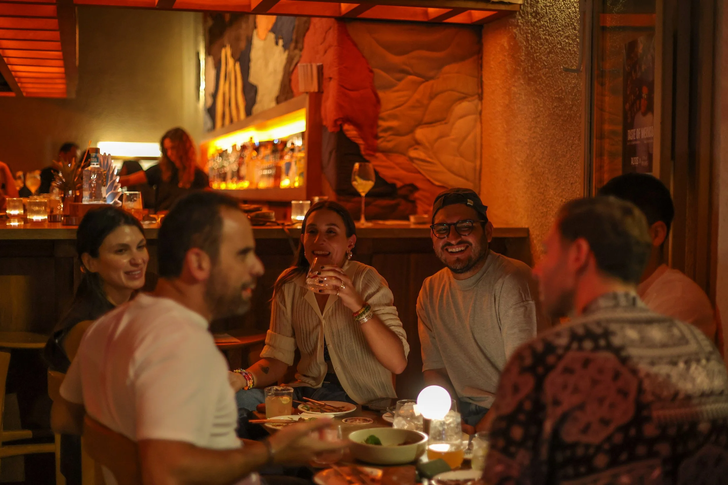 A group of friends sitting at a table in a dimly lit restaurant or bar, enjoying drinks and conversation, with a bar area and colorful artwork visible in the background.