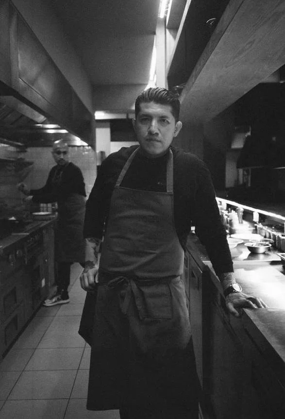 Young man wearing a black shirt and apron standing in a modern kitchen, with another chef in the background cooking.