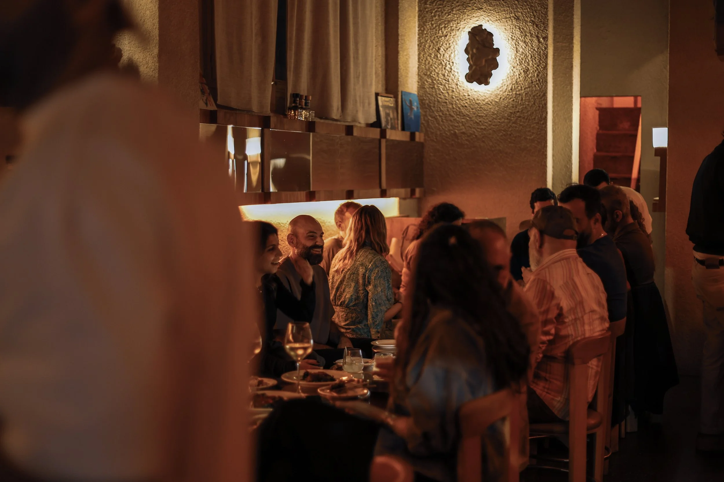 People gathered at a dimly lit restaurant, sitting at a long table, socializing and enjoying drinks.