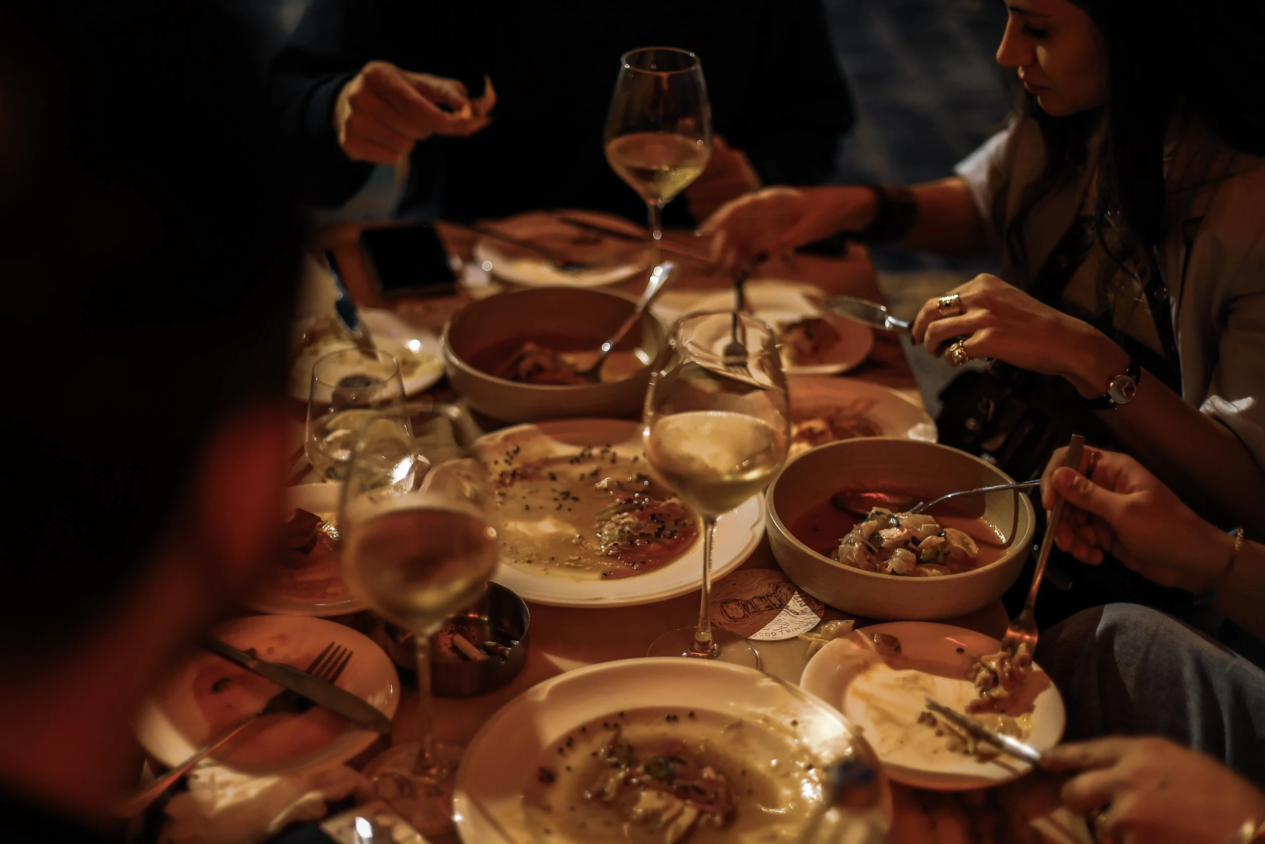 People dining at a restaurant table with wine glasses, plates, and bowls of food, enjoying a meal together in a dark, cozy setting.