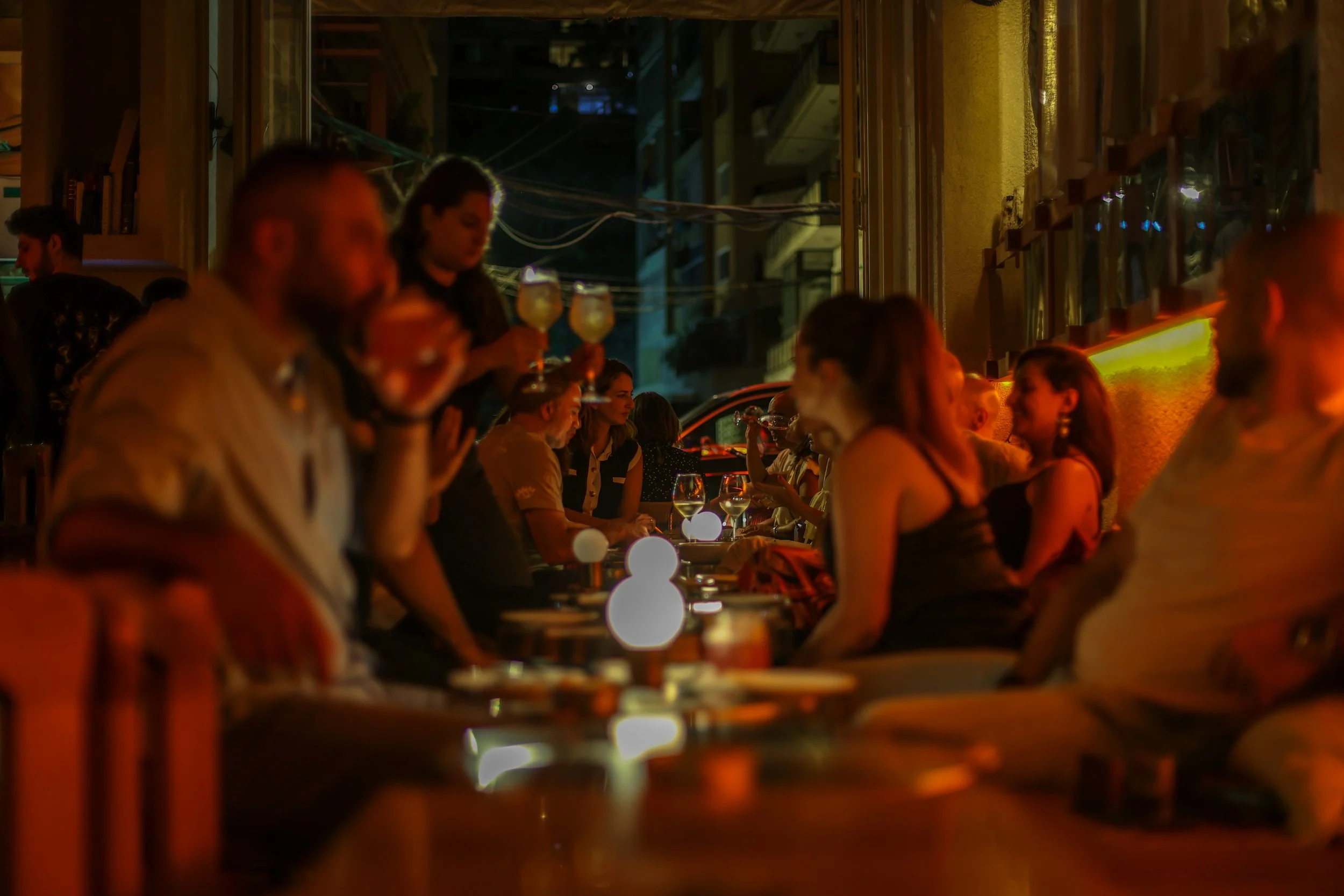 People socializing at a dimly lit restaurant or bar, with some holding drinks and sitting at tables along a window, creating a lively, cozy atmosphere.