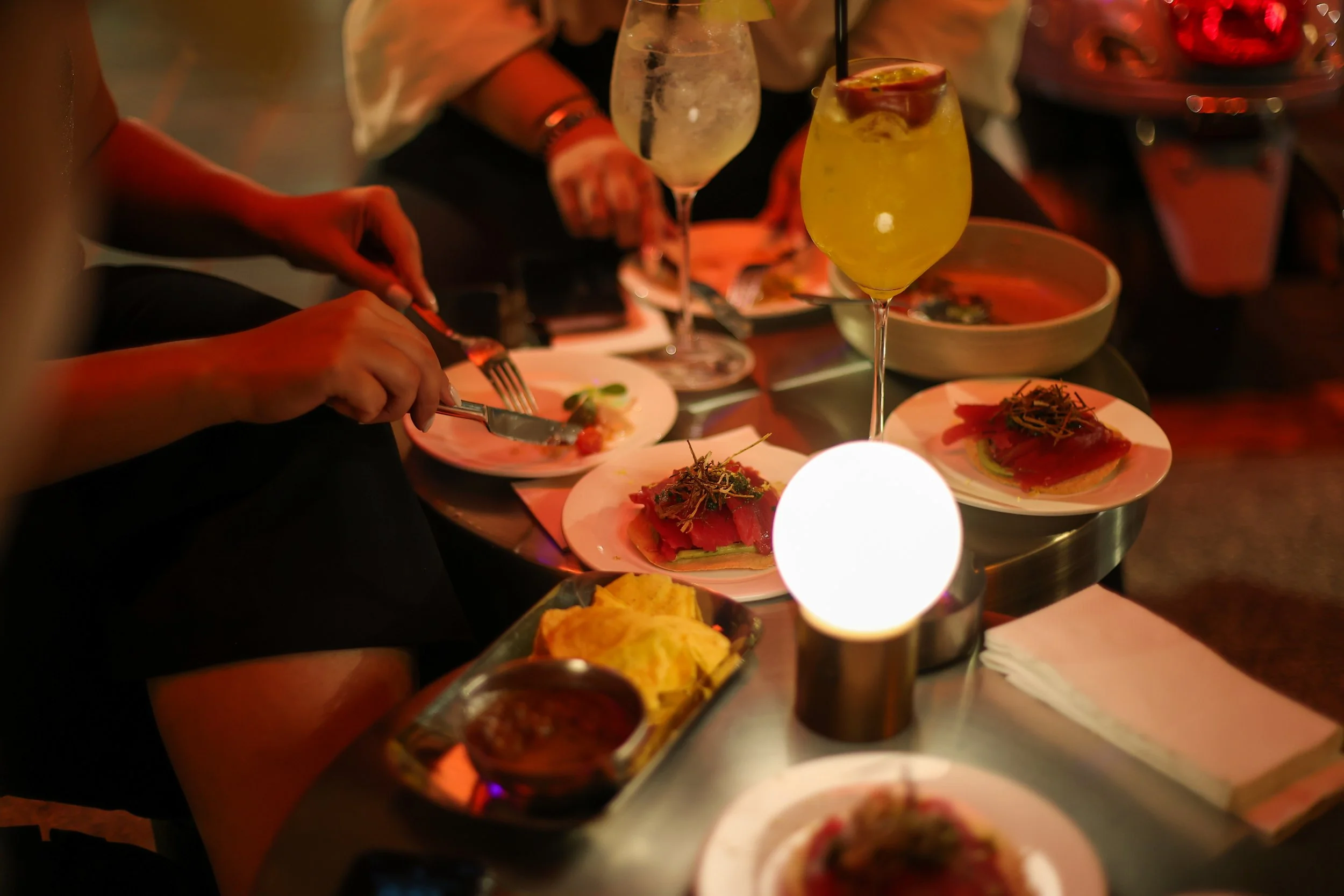 A dinner table with plates of sashimi, cocktails, chips, and a small dish of sauce, illuminated by a warm glowing light in a dimly lit setting.