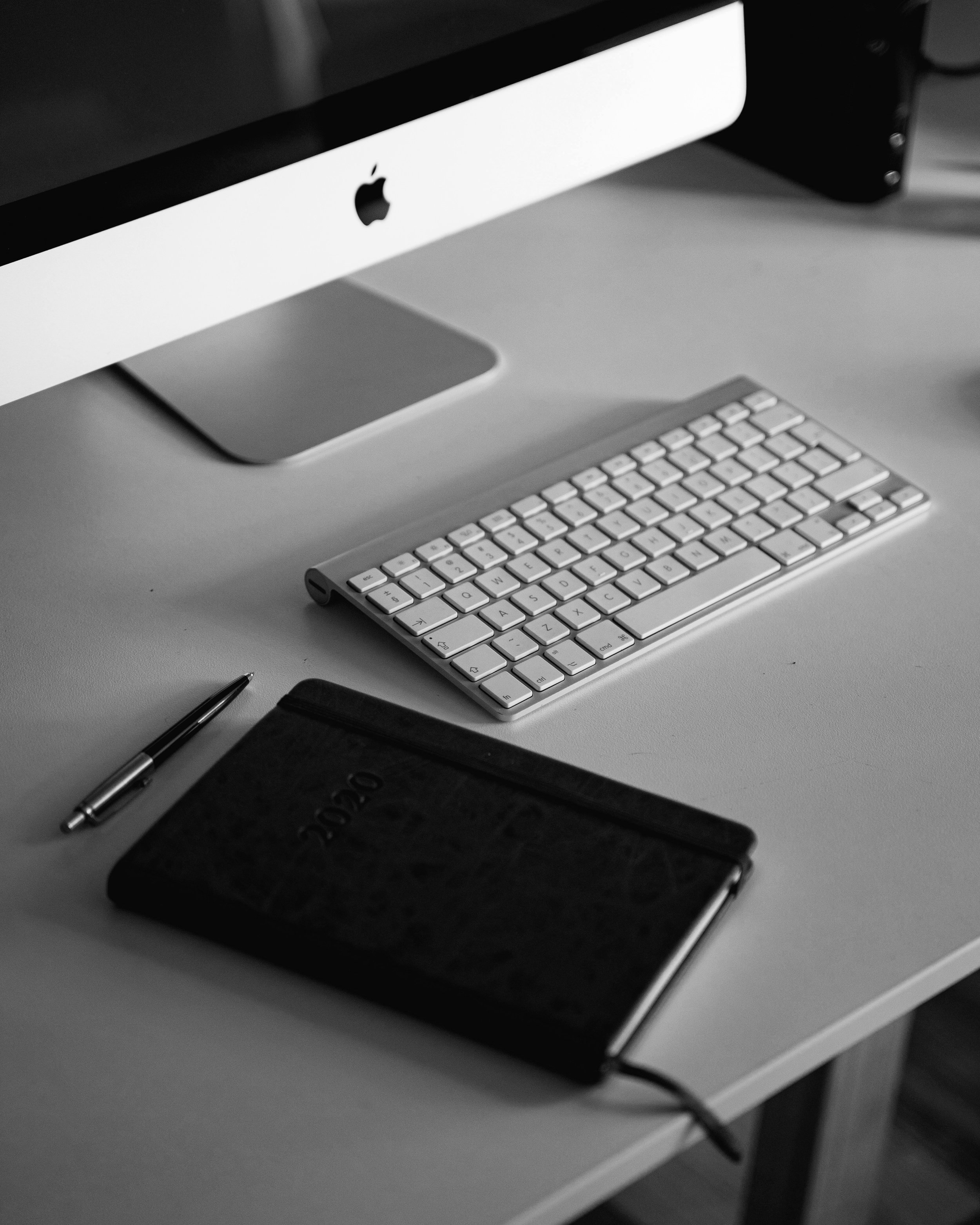 Black and white photo of an iMac computer, a wireless keyboard, a pen, and a closed notebook on a desk.