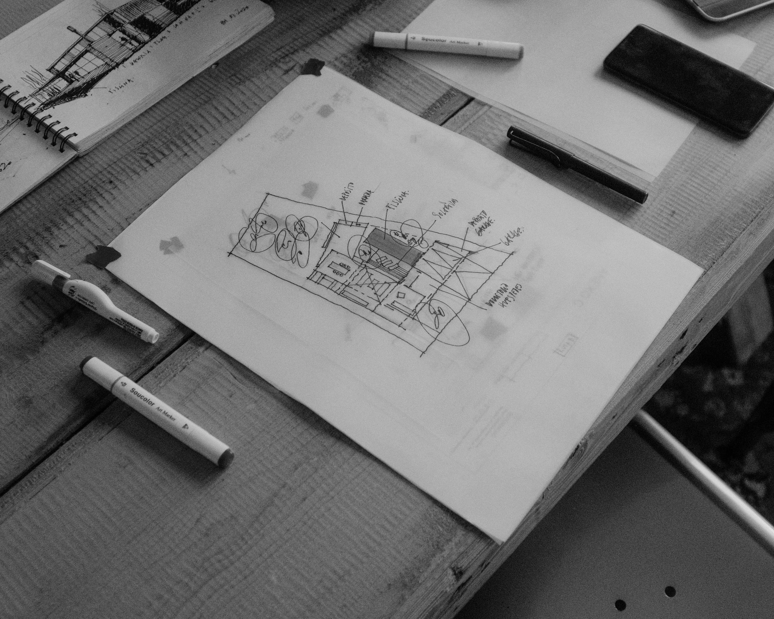 A desk with architectural sketches and plans, black and white, including a hand-drawn building layout, pens, markers, a smartphone, and paper.