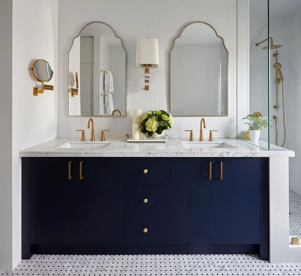A modern bathroom vanity with a navy blue cabinet, a white marble countertop, dual sinks with gold faucets, and two large mirrors. The mirror area has decorative wall-mounted mirrors and a white wall sconce with gold accents. There are plants and can