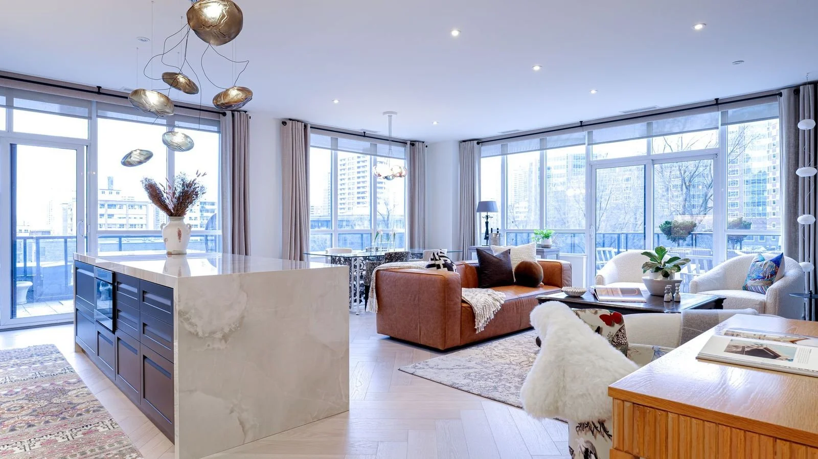 Bright open-concept living room with floor-to-ceiling windows, modern furnishings, a brown sofa with pillows, white chairs, plants, and cityscape view.