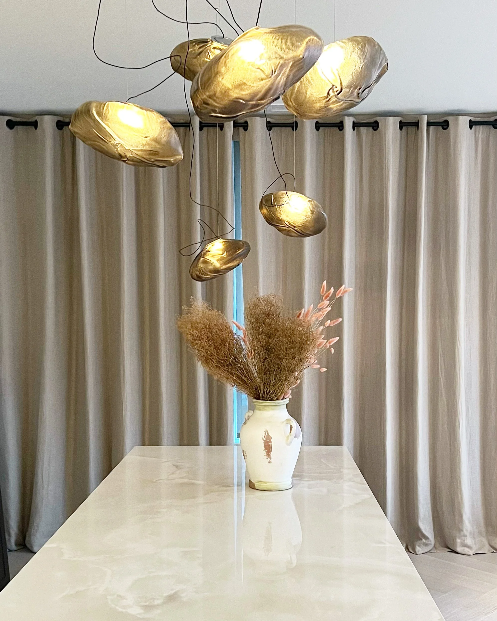 Vase with dried flowers on a white table, hanging gold sculpture lights above, beige curtains in the background.