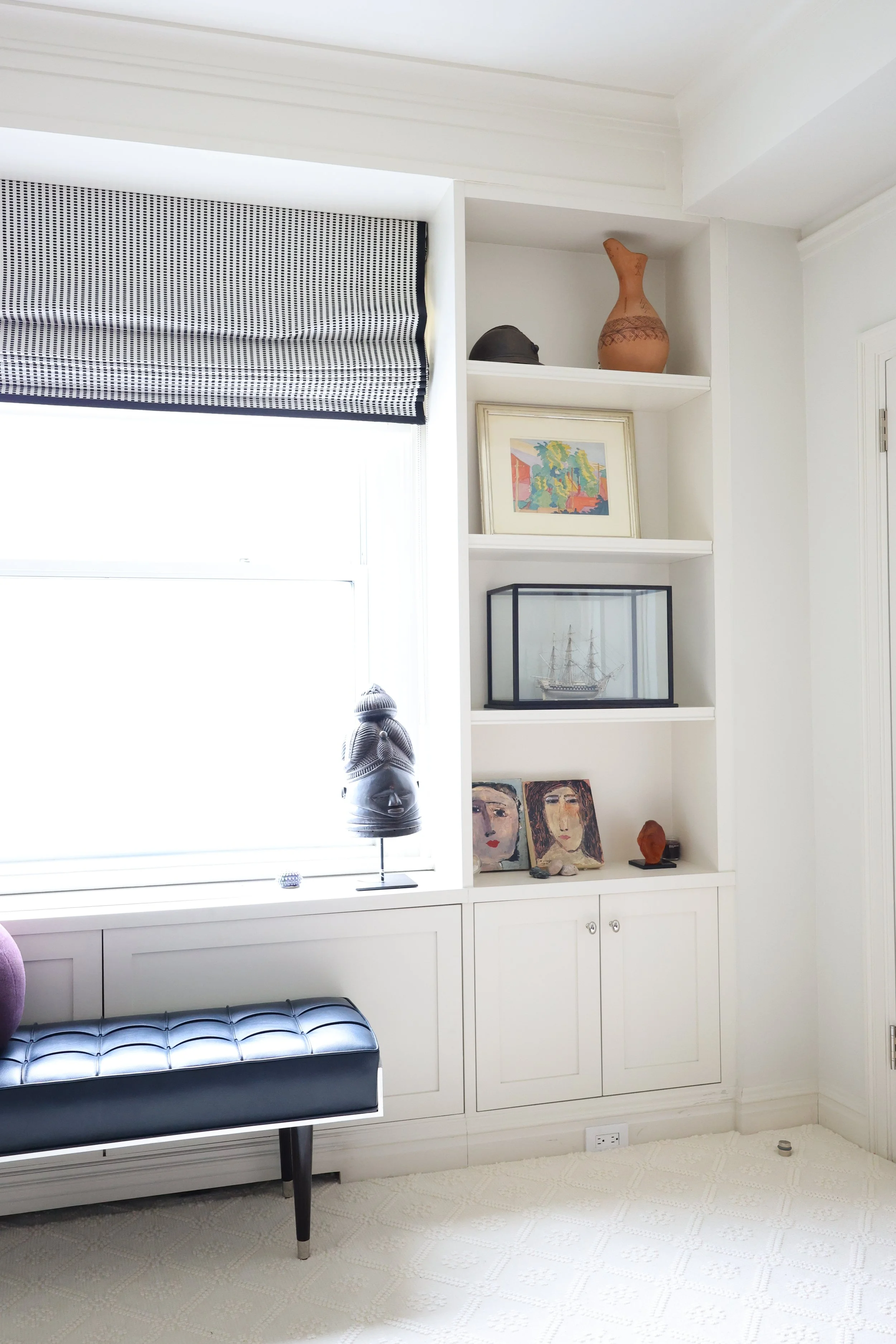 Interior of a room with a white built-in cabinet and shelves, a bench with black upholstery, a window with a black and white patterned Roman shade, decorative objects, paintings, and a model ship displayed on the shelves.