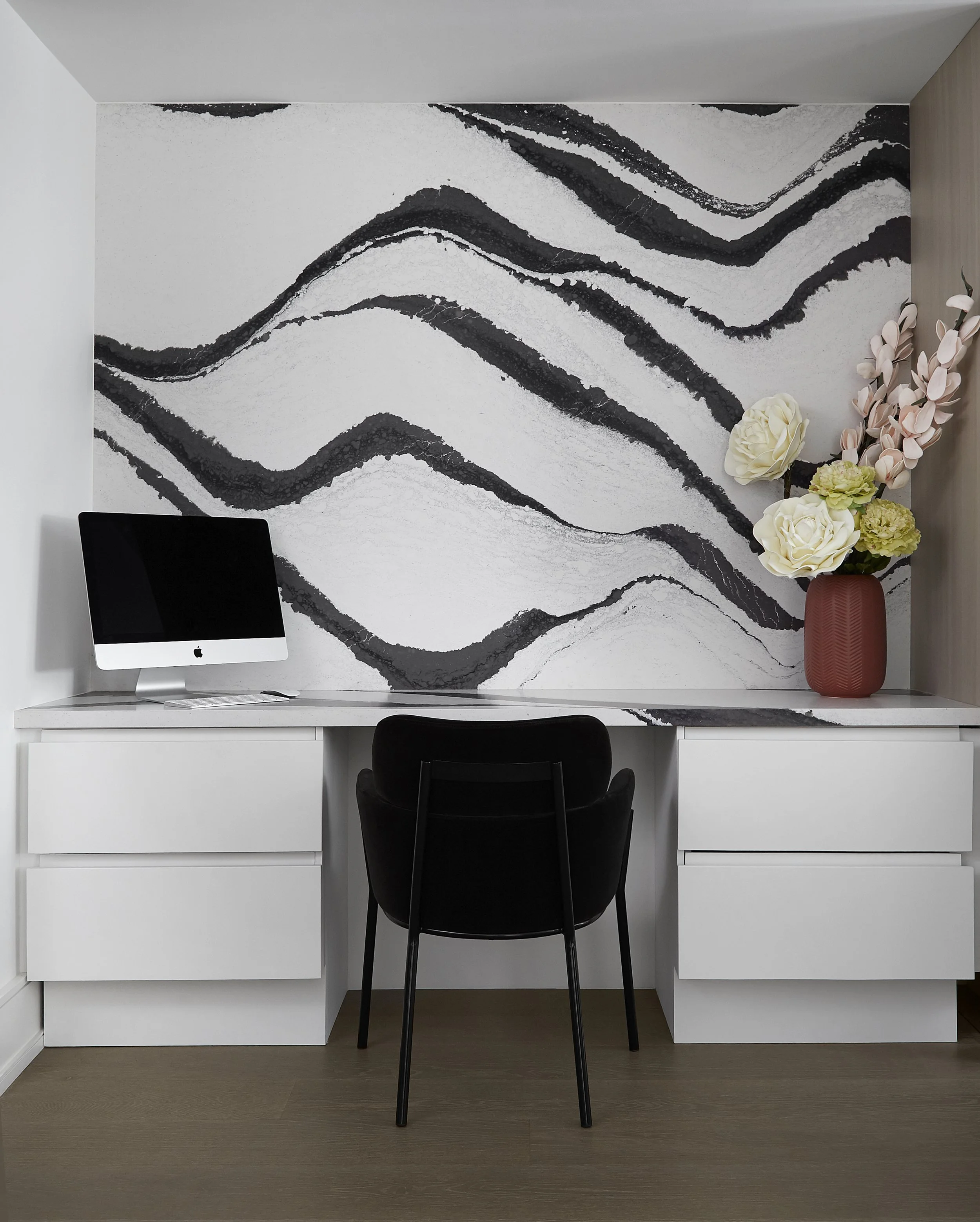 Home office desk with an iMac, black chair, and a flower vase with large white and pink flowers in front of a black and white abstract wall mural.