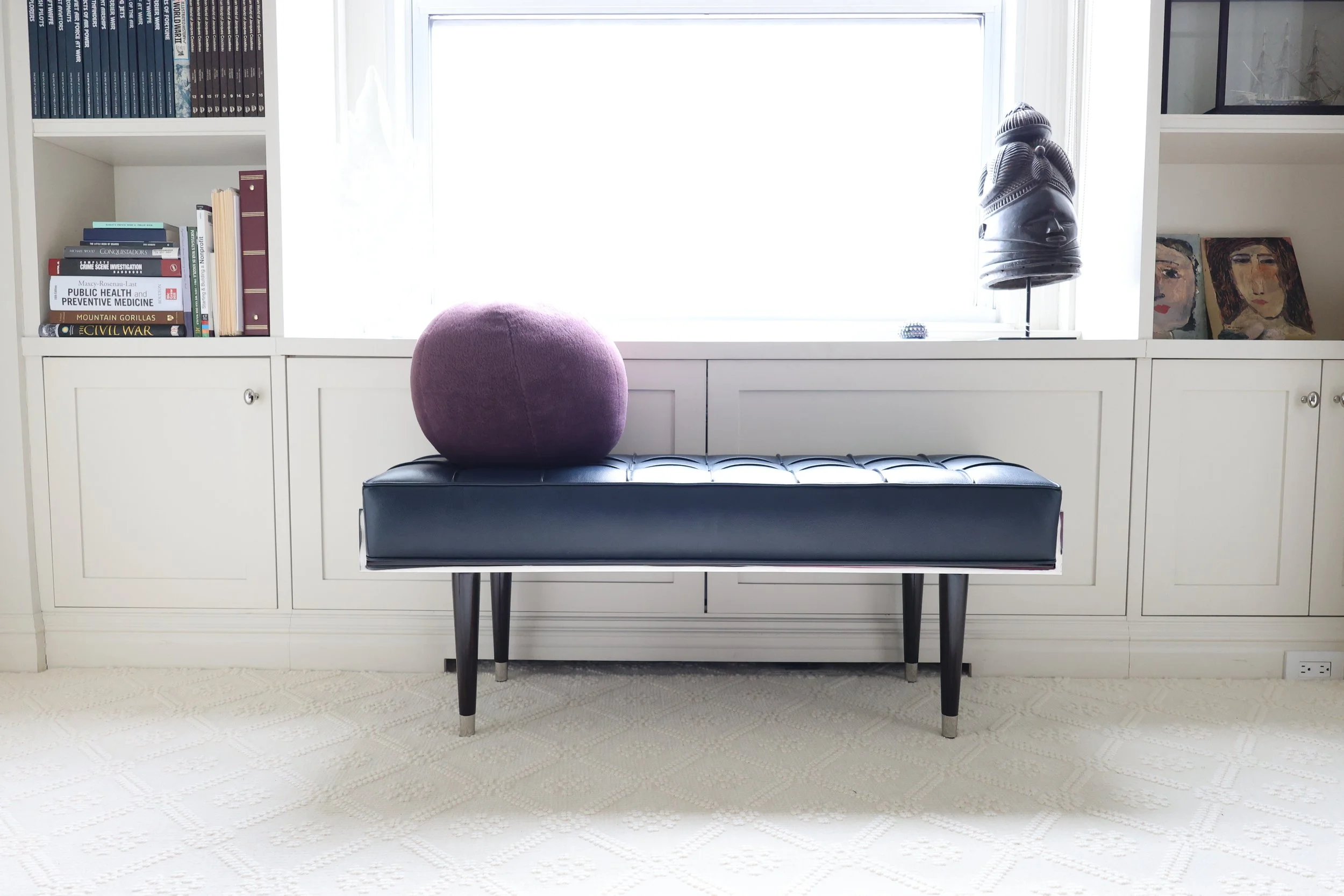 Interior view of a room with a black upholstered bench topped with a purple round pillow, against a white cabinet with books, paintings, and a tribal mask on the shelves, and a window with sunlight in the background.
