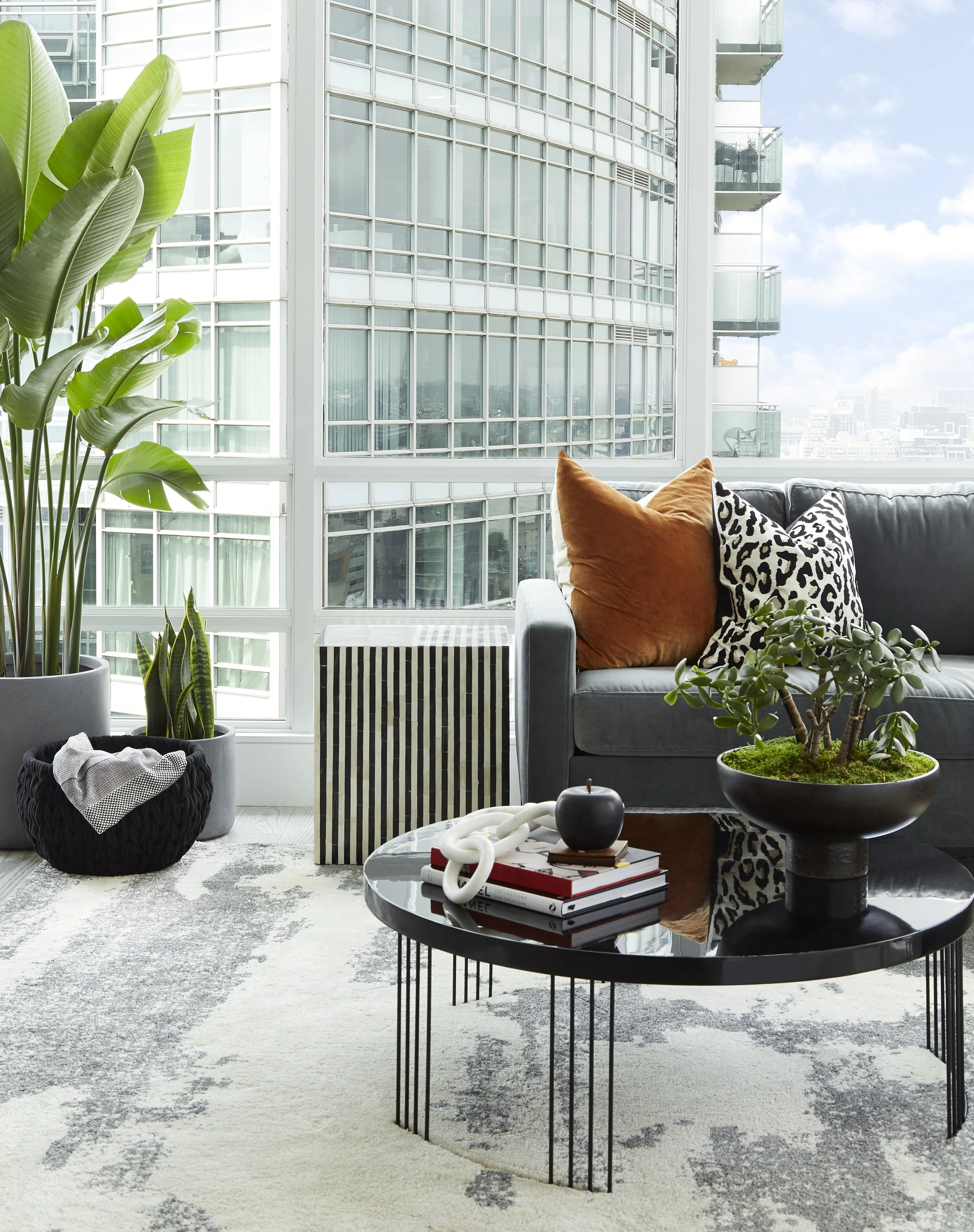 Modern living room with plants, black coffee table with decorative items, gray sofa with colorful pillows, and city skyline view.