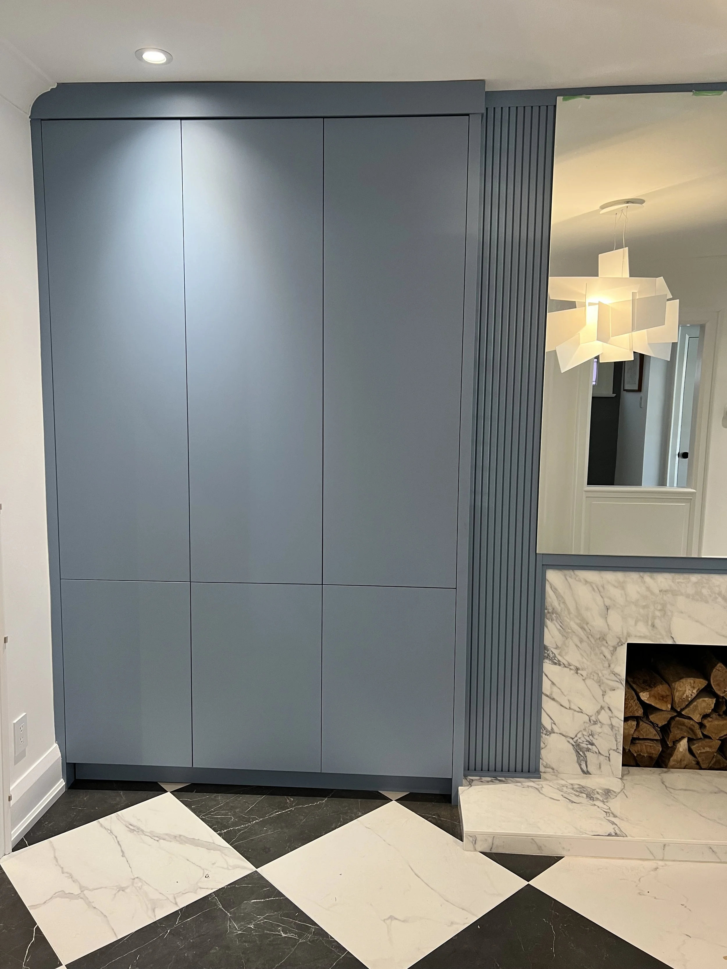 Blue built-in cabinet next to a marble fireplace with wood logs inside, black and white checkered marble floor, modern pendant light in the background.