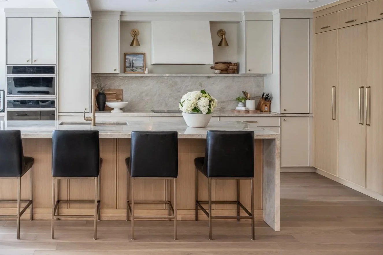Modern kitchen with light wood and white cabinetry, black barstools at a marble island, decorated with a white flower vase, and minimal decor.