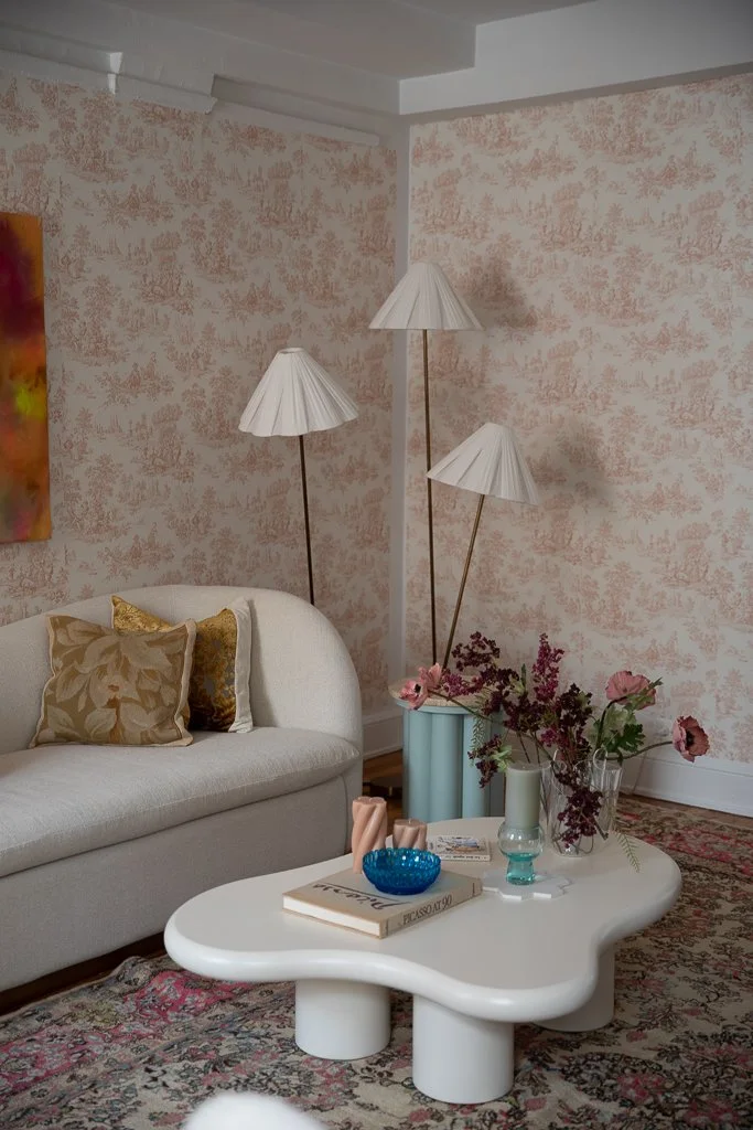 Living room with a white sofa with gold and brown pillows, a unique white coffee table with curved edges, tall floor lamps with white pleated lampshades, a blue vase with pink flowers, a small turquoise candle holder, a book, and a patterned area rug