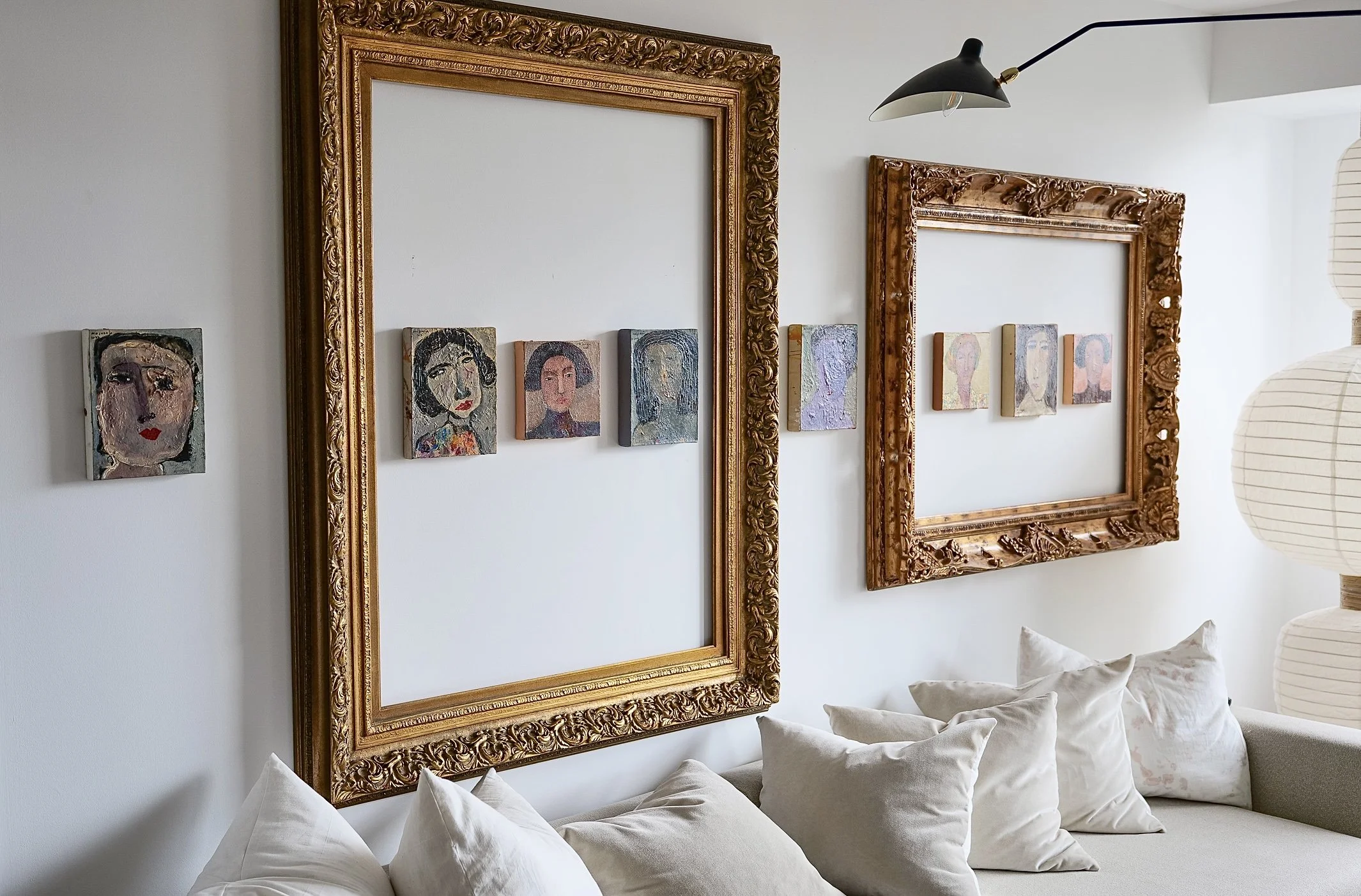 Living room with ornate gold picture frames on white wall, small colorful portraits inside frames, white couch with multiple white pillows, paper lanterns on right side, ceiling light fixture above.