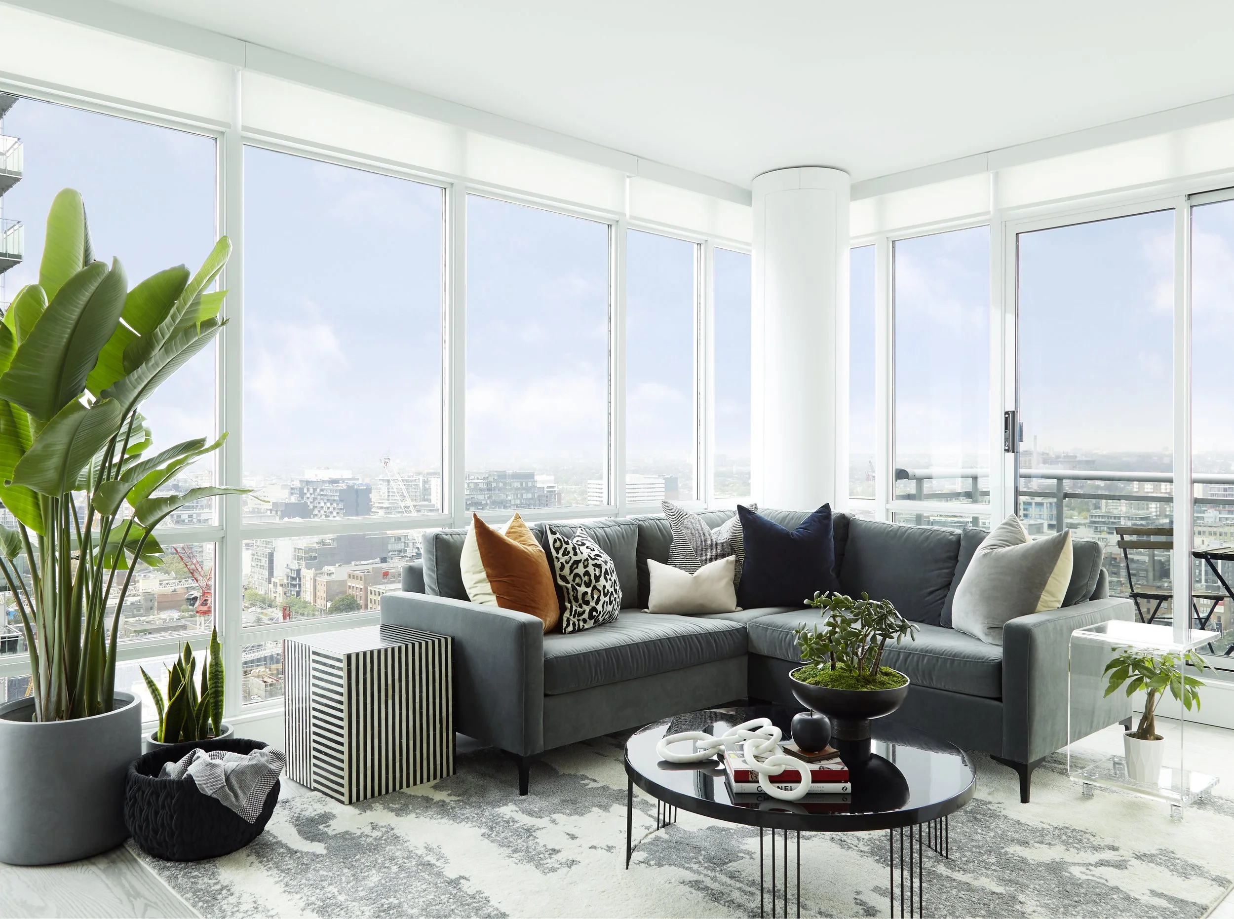 Modern living room with large floor-to-ceiling windows, gray sectional sofa with colorful pillows, black round coffee table with decorative items, large indoor plants, and city skyline views