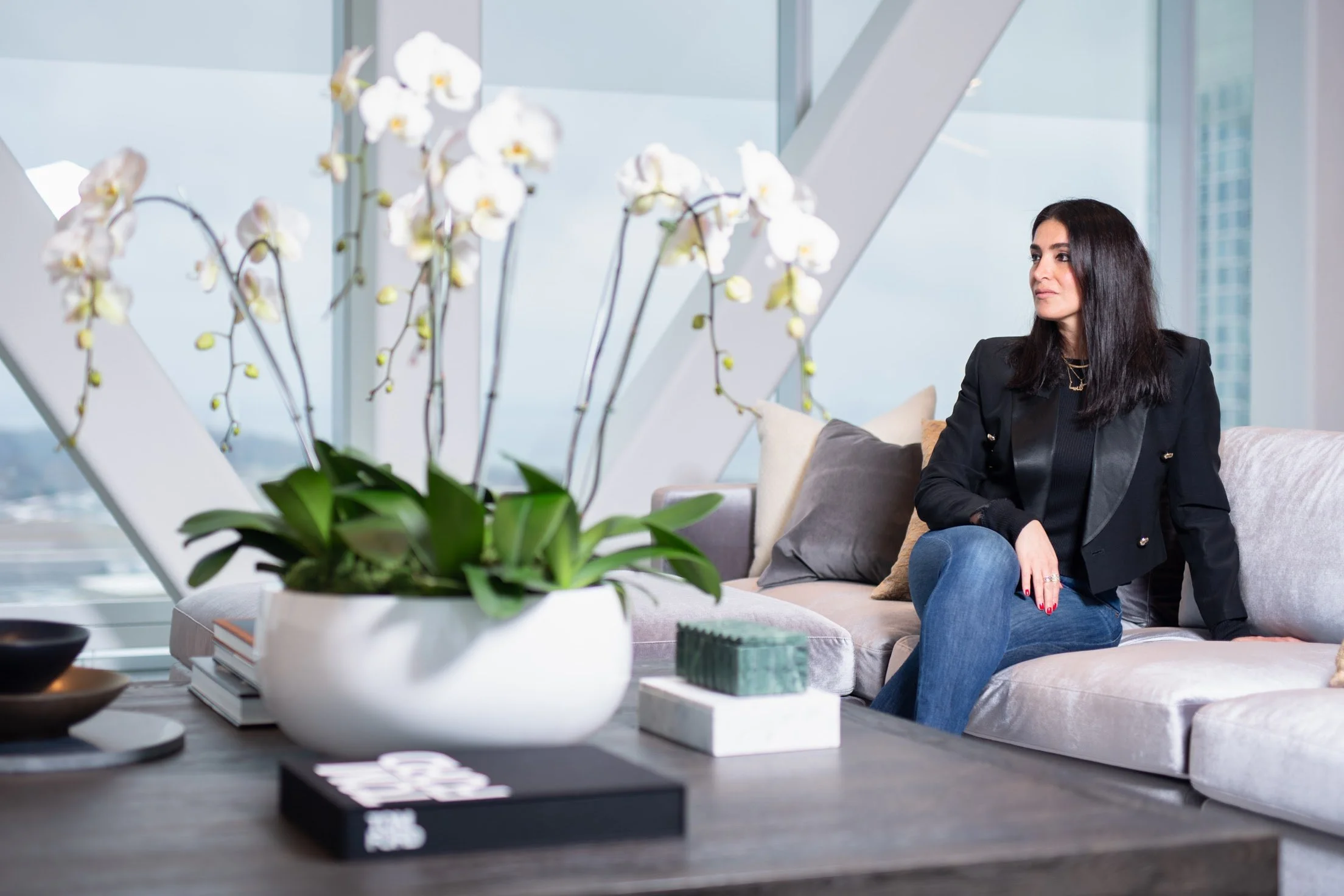 Woman with black hair sitting on a beige sofa in a modern apartment with floor-to-ceiling windows, next to a table with a large white orchid plant, books, and decorative items.