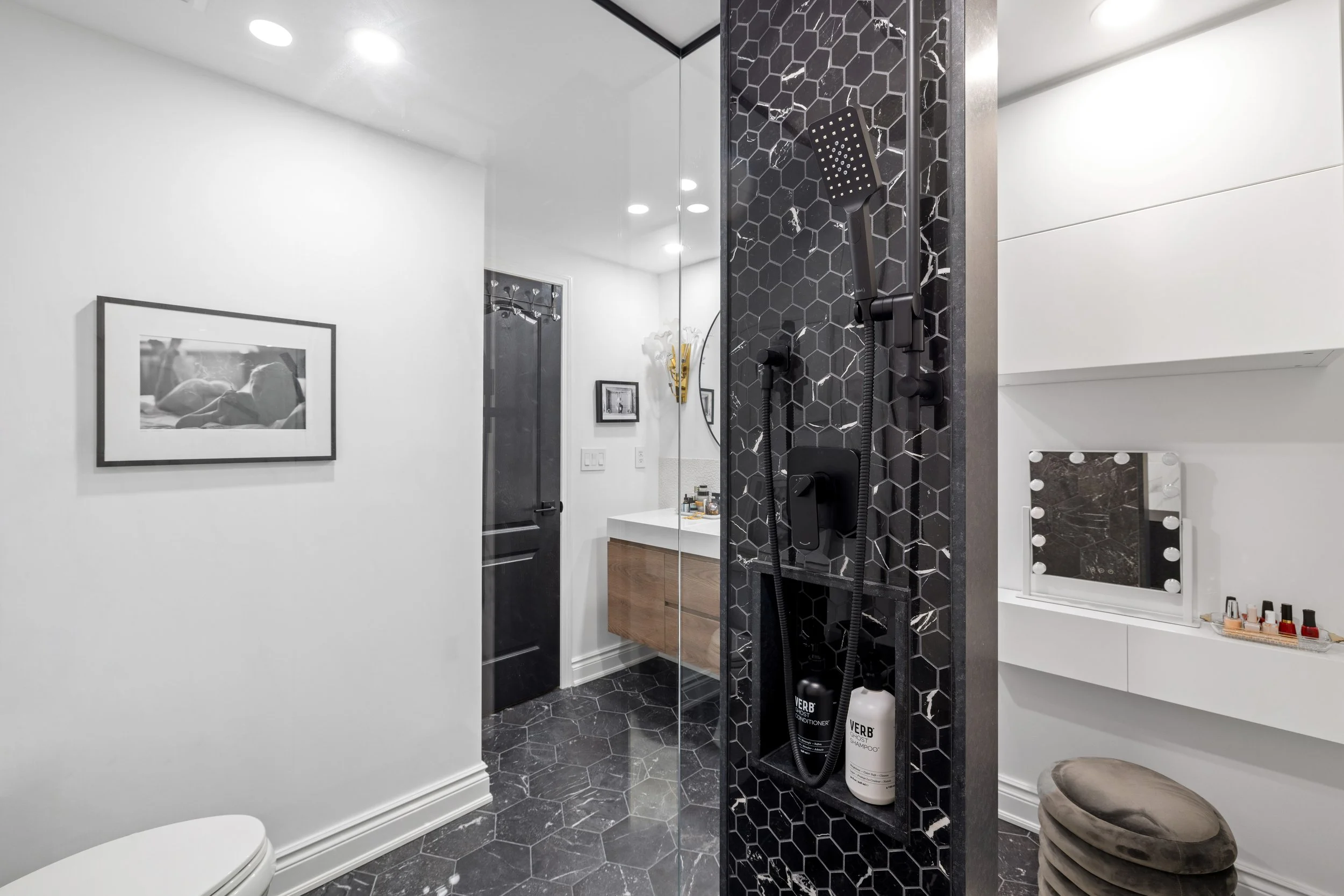 Modern bathroom with black hexagon tile floor and walk-in shower featuring black hexagon tile walls and a black shower head. A white vanity with a mirror and skincare products, a black door, and minimalist white walls with a black-framed photograph.
