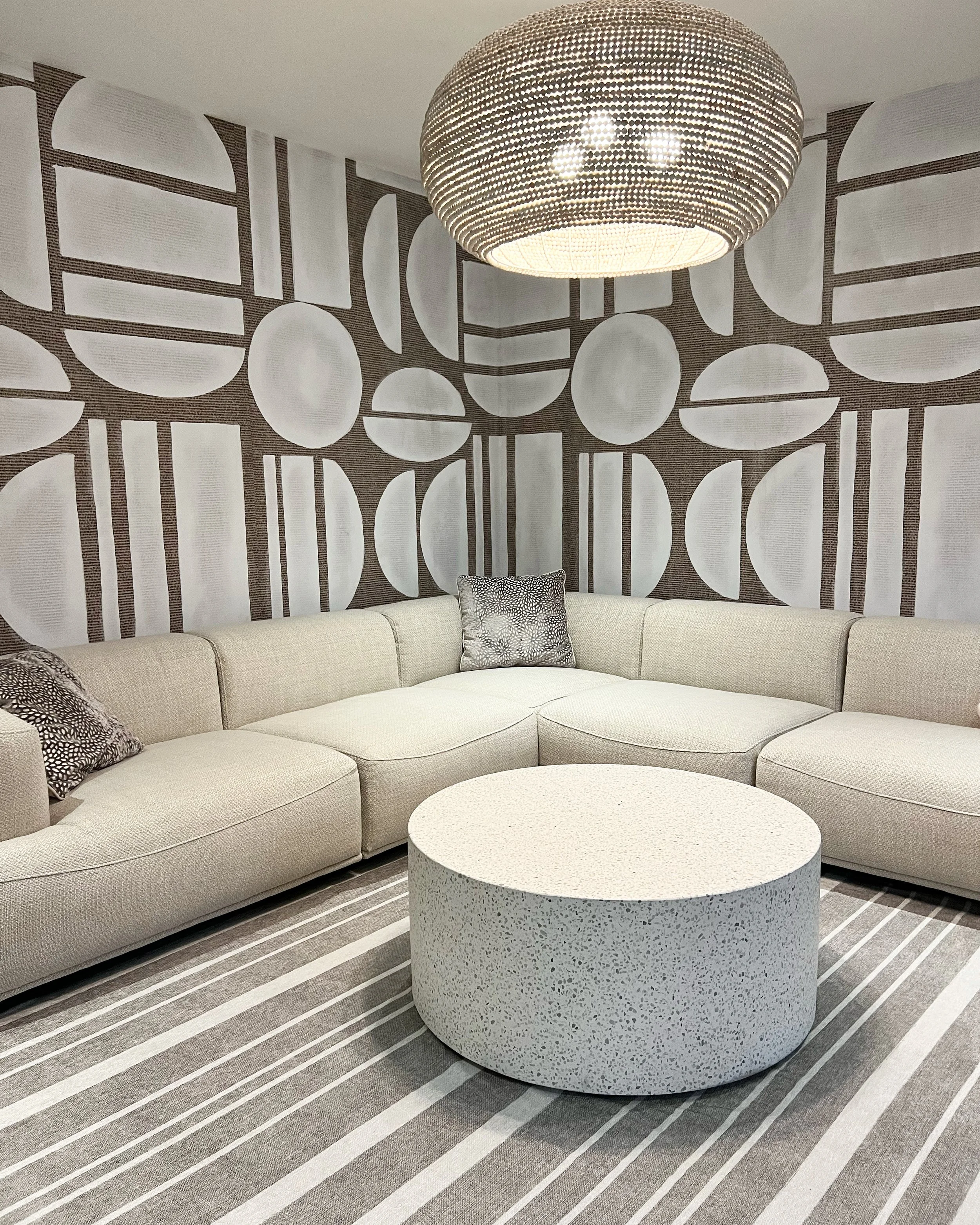 Modern living room with a beige sectional sofa, decorative pillows, a circular coffee table, geometric wallpaper, and a round ceiling light fixture.