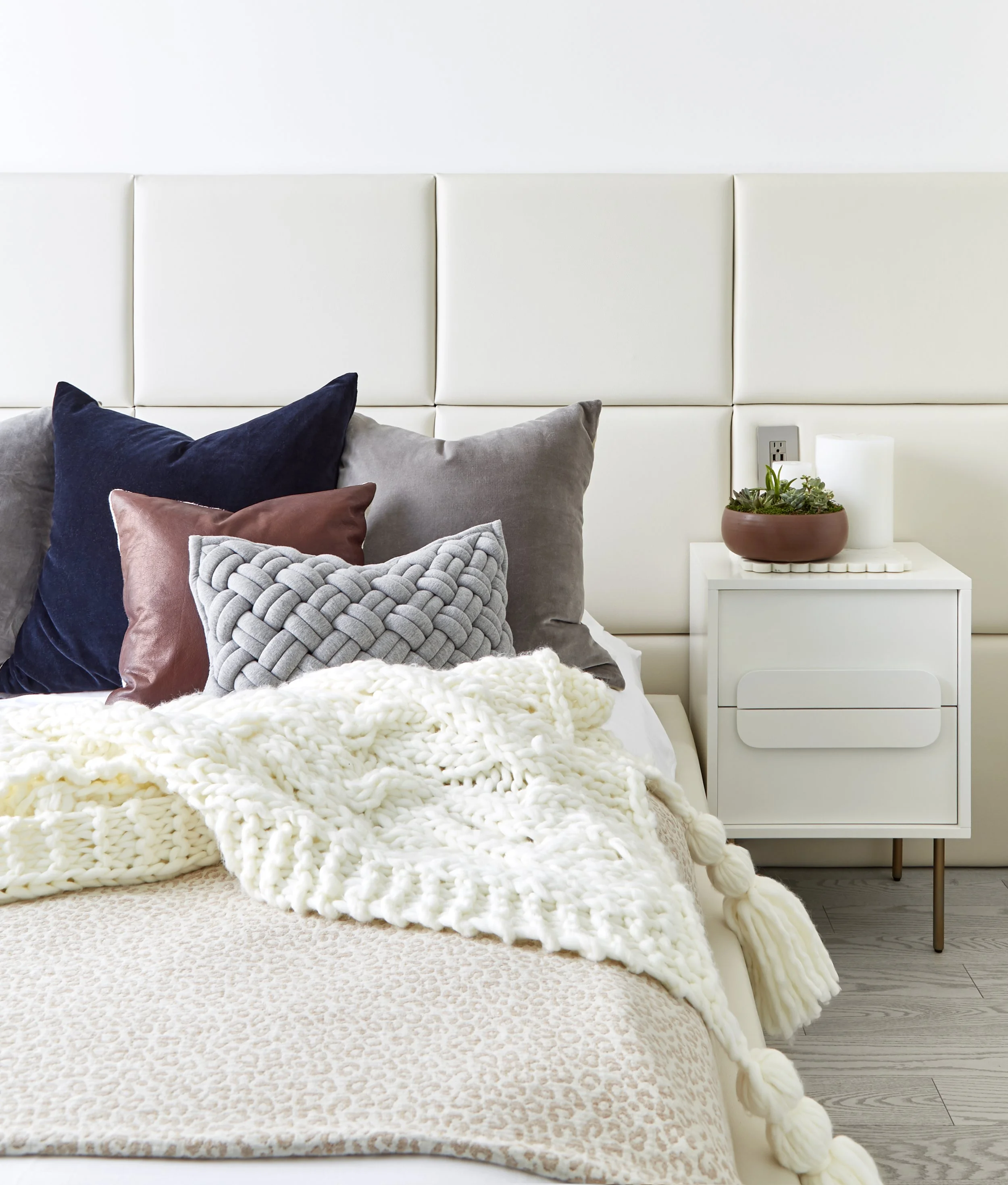 Modern bedroom with a white padded headboard, a variety of pillows including navy, brown, gray, and a knitted gray pillow, a cream chunky knit blanket, and a beige bedspread with a subtle animal print. A white nightstand holds a round brown planter w