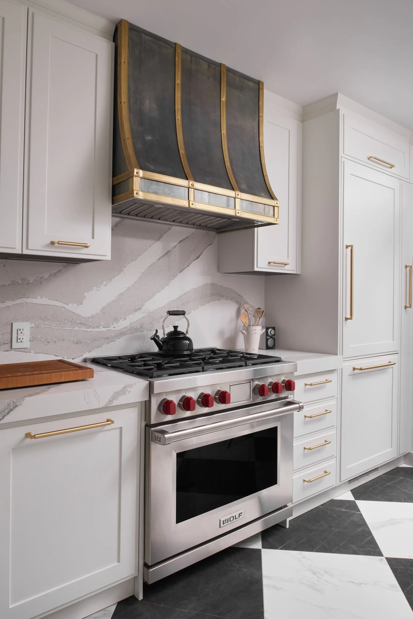 Modern kitchen with white cabinets, a stainless steel wolf oven with red knobs, a black teapot on the stove, and a decorative gold and black range hood. The floor has black and white tiles.