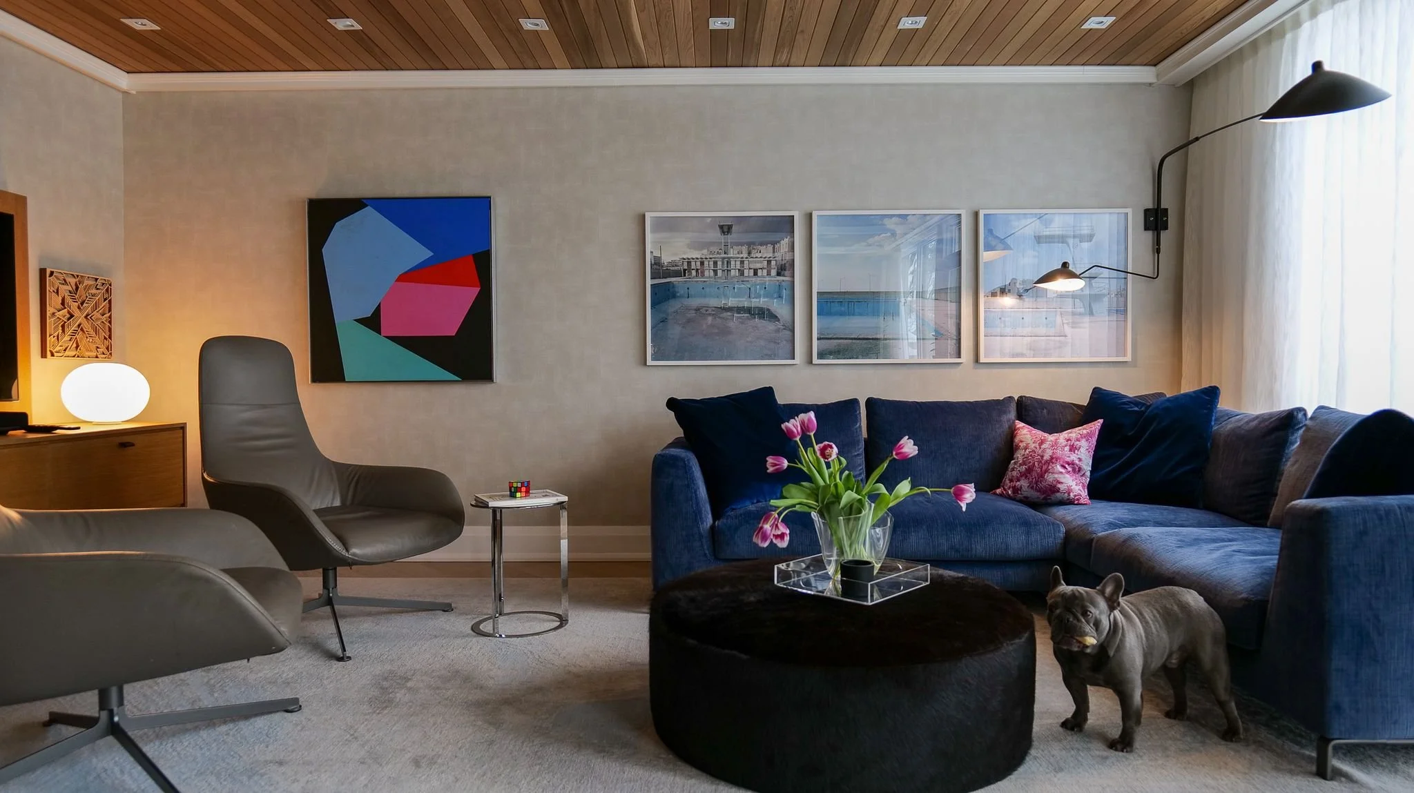 Living room with a blue sectional sofa, pink tulips in a glass vase on a round black ottoman, French bulldog, wall art including four landscape photos, and a corner lamp near a window with sheer curtains.