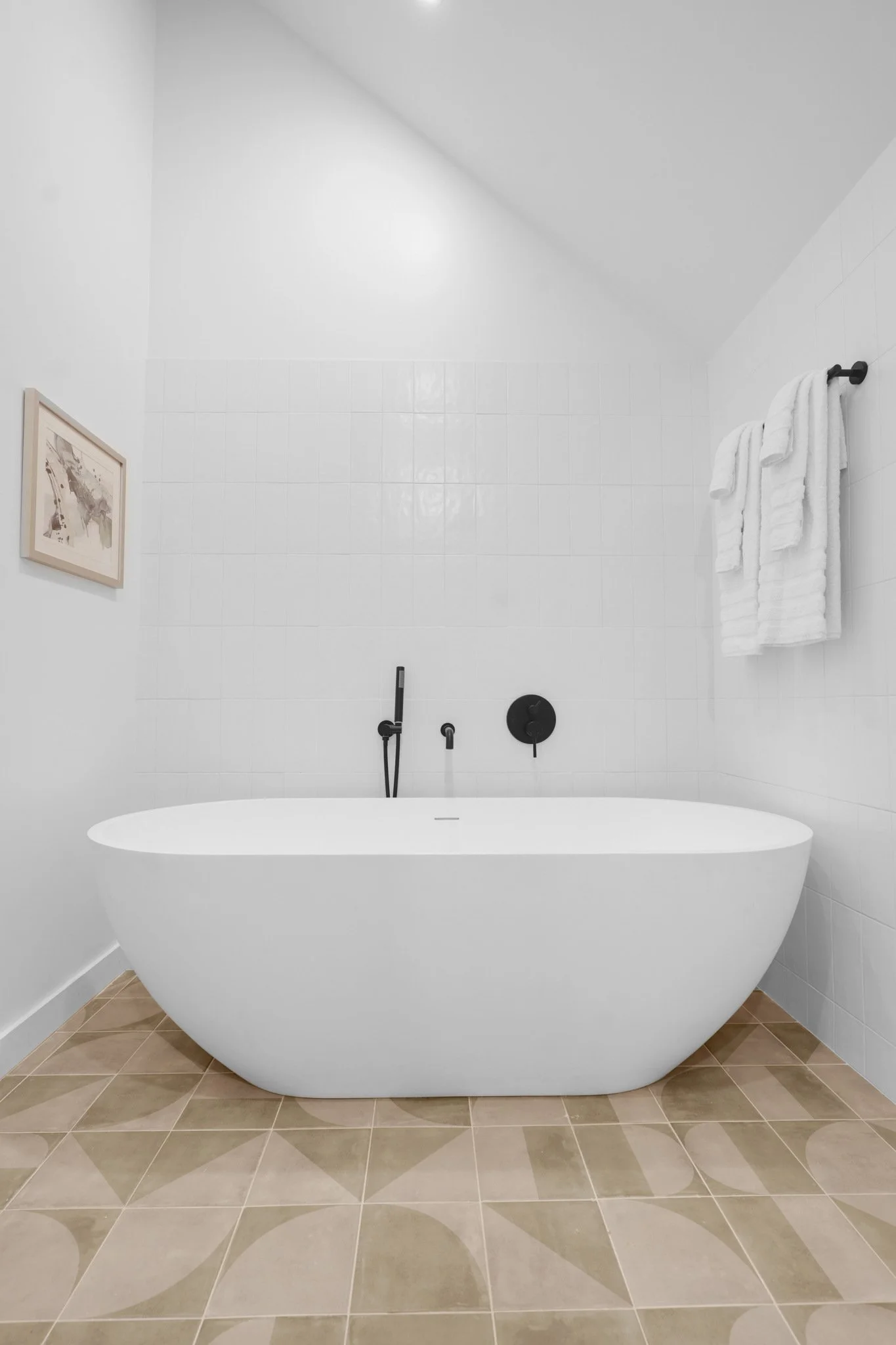 Modern white freestanding bathtub in a minimalist bathroom with white tiled walls, beige patterned floor tiles, black fixtures, and a towel rack with white towels.
