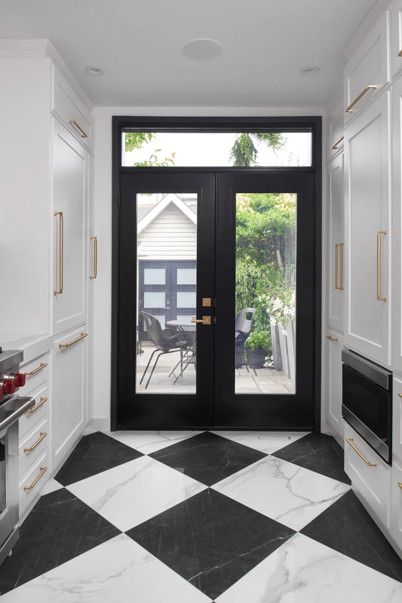 Modern kitchen with black-framed glass doors leading to a patio, white cabinets with gold handles, black and white checkered marble floor.