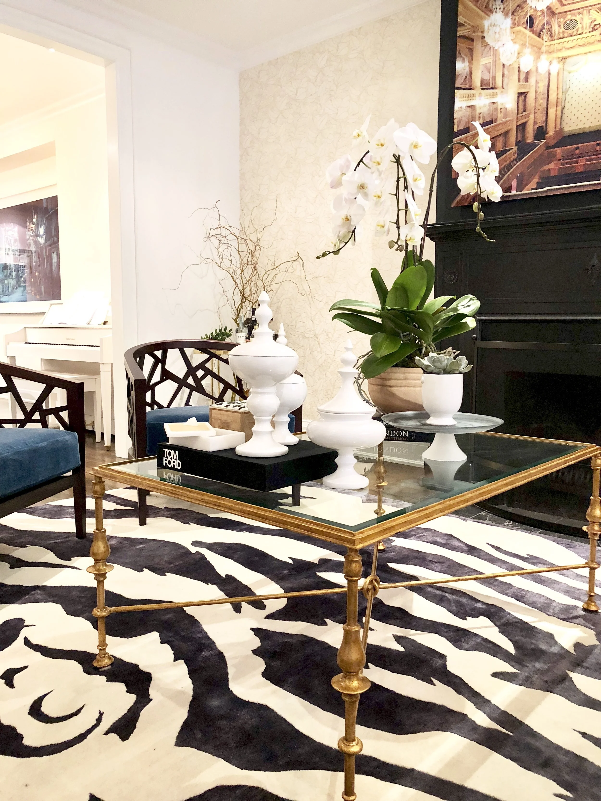 Living room with a gold and glass coffee table, white vases with decorative branches, orchids, and succulents, a zebra print rug, and a black fireplace mantle with a large framed picture on it.