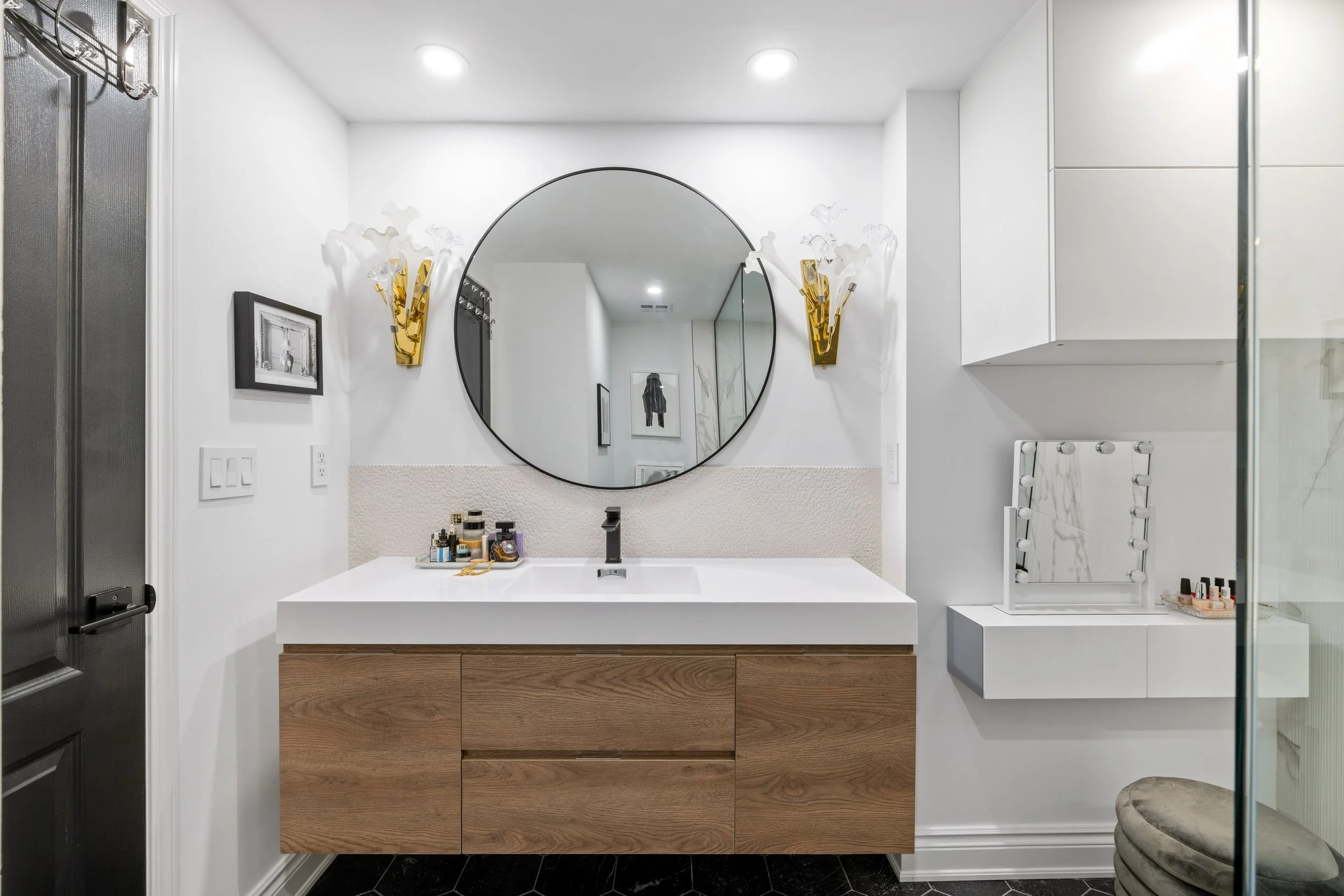 Modern bathroom with white walls, a round mirror above a wooden vanity with a white countertop, gold wall sconces with white flower-shaped shades, a small framed photograph, and a vanity mirror with lights on a white cabinet. A black door is partiall
