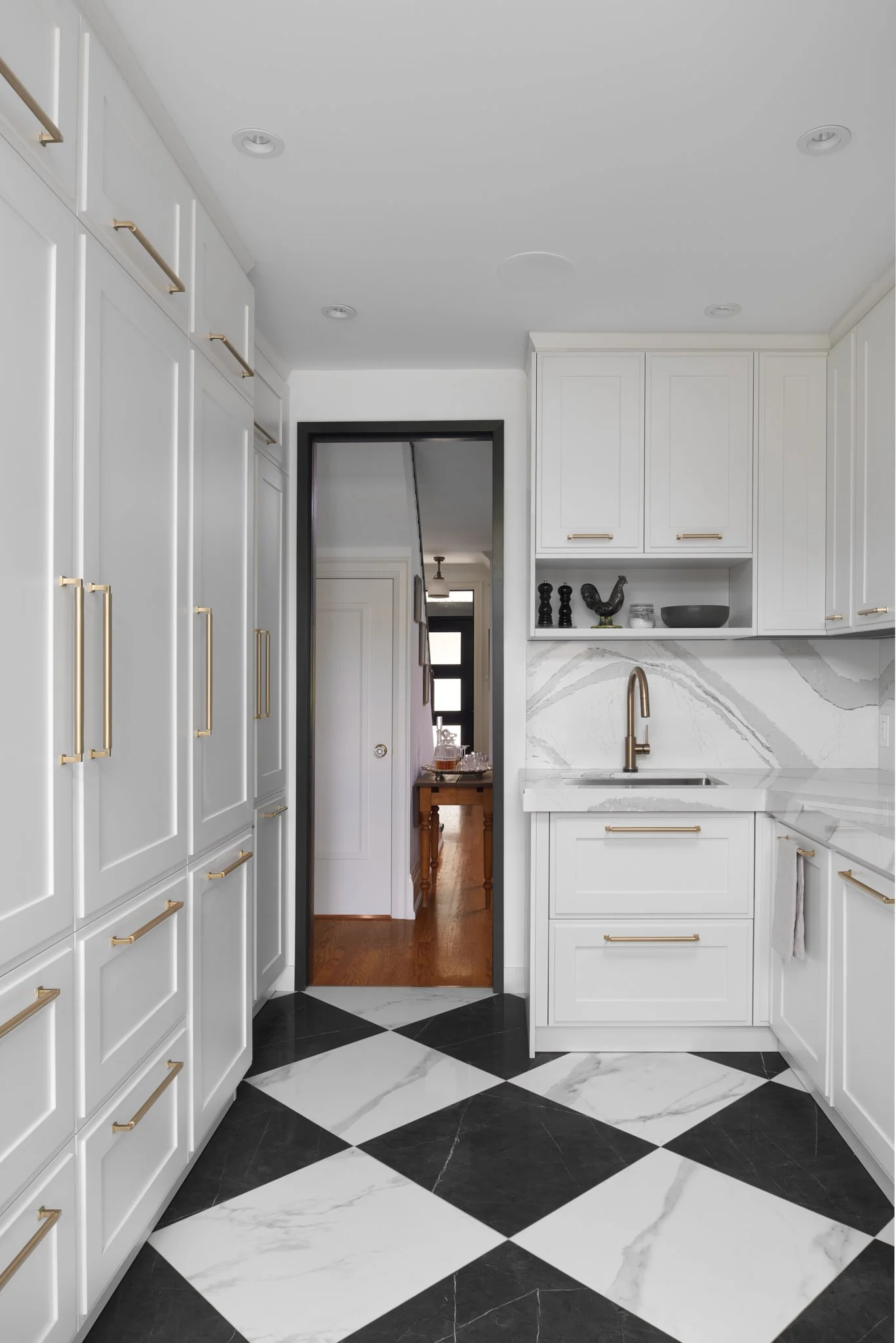 Modern kitchen with white cabinets and gold handles, black and white diamond-patterned marble floor, view looking toward a doorway with a dark frame, revealing a hallway with wooden flooring and black front door.