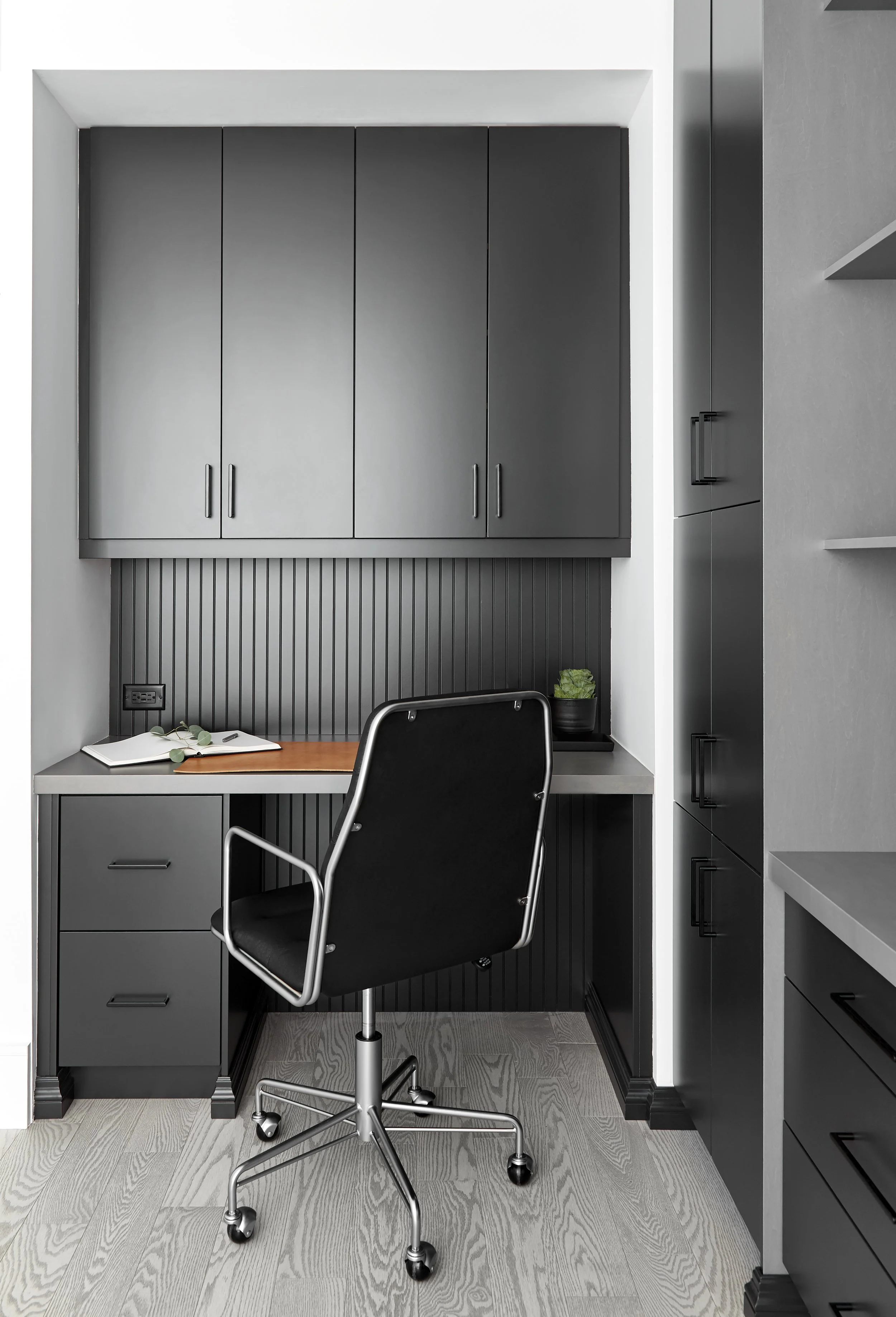 Modern home office nook with gray cabinetry, black beadboard accent wall, black swivel chair, and minimalist decor including a small potted plant and open notebook.