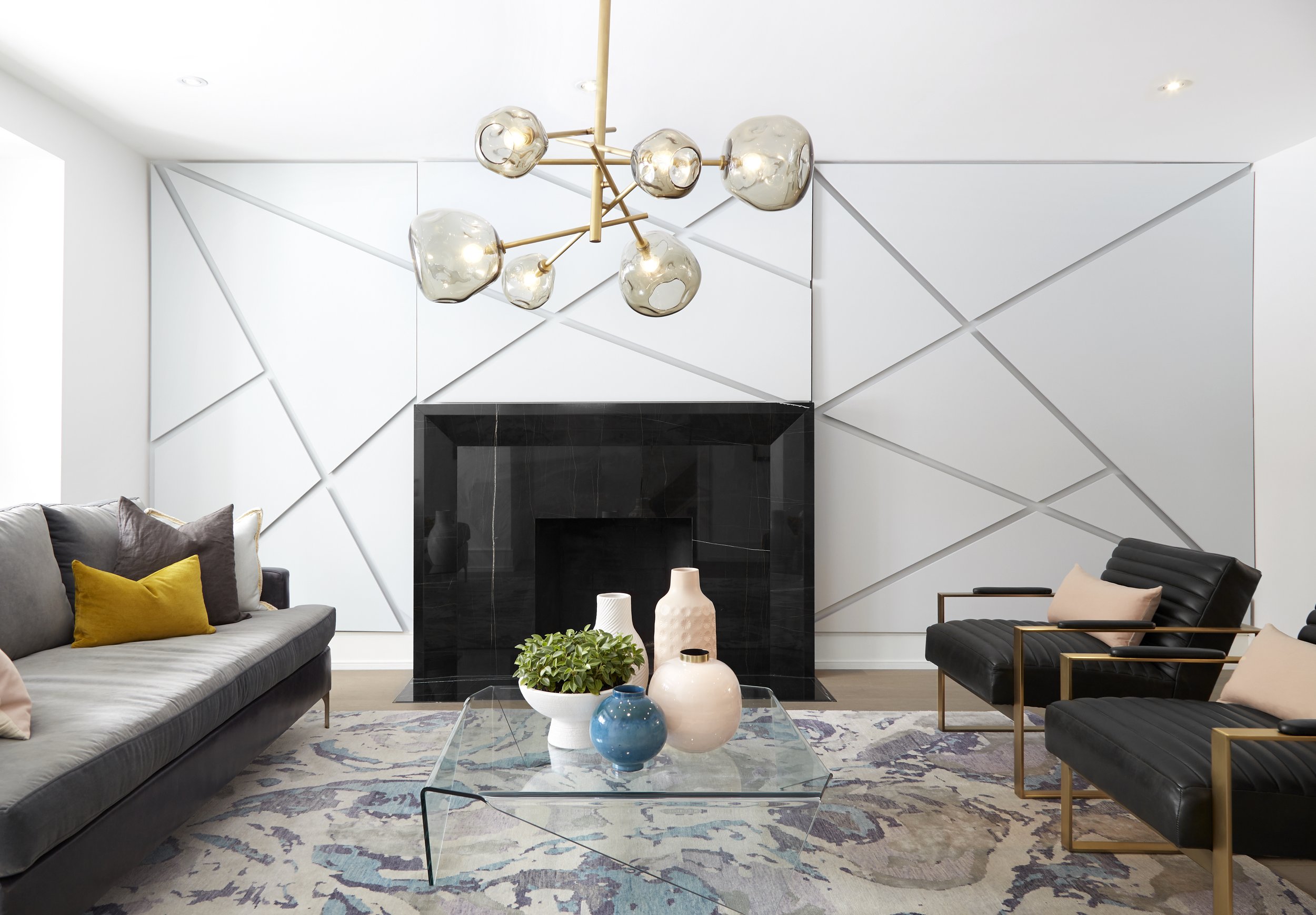 Modern living room with a white geometric wall design, a black fireplace, and a glass coffee table with vases and a plant. There are sofas with pillows, one in grey and two in black with gold frames.