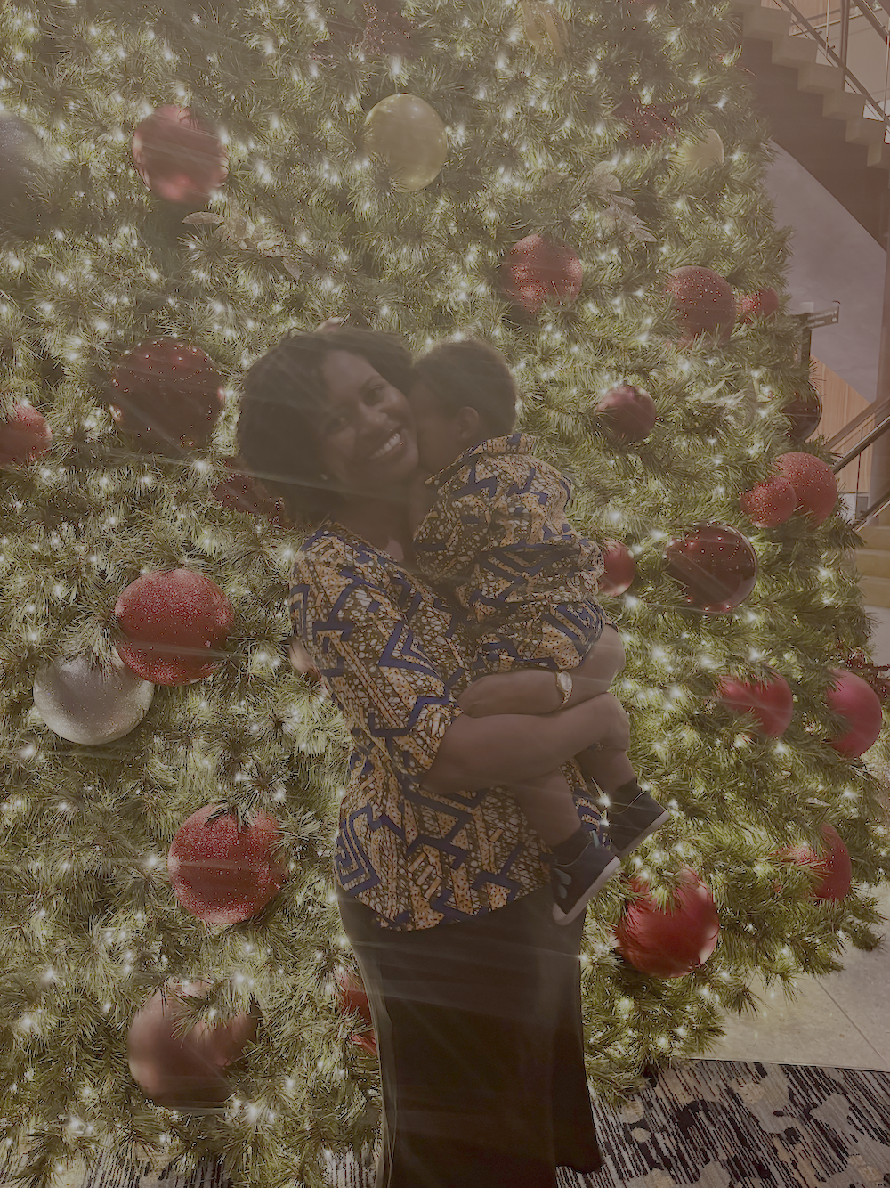 A woman holding a child in front of a decorated Christmas tree with red and gold ornaments, illuminated with warm yellow lights.