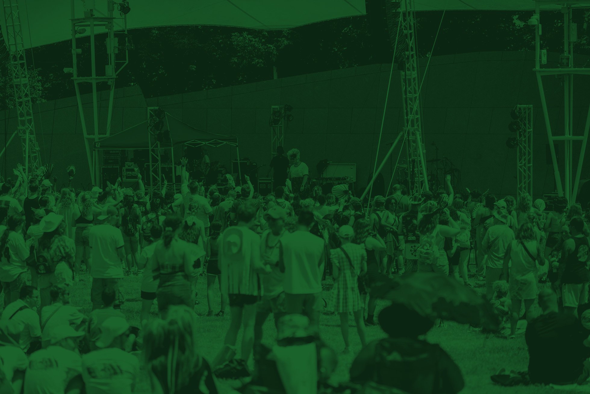 Crowd of people gathered outdoors at a concert or festival with a stage and sound equipment.