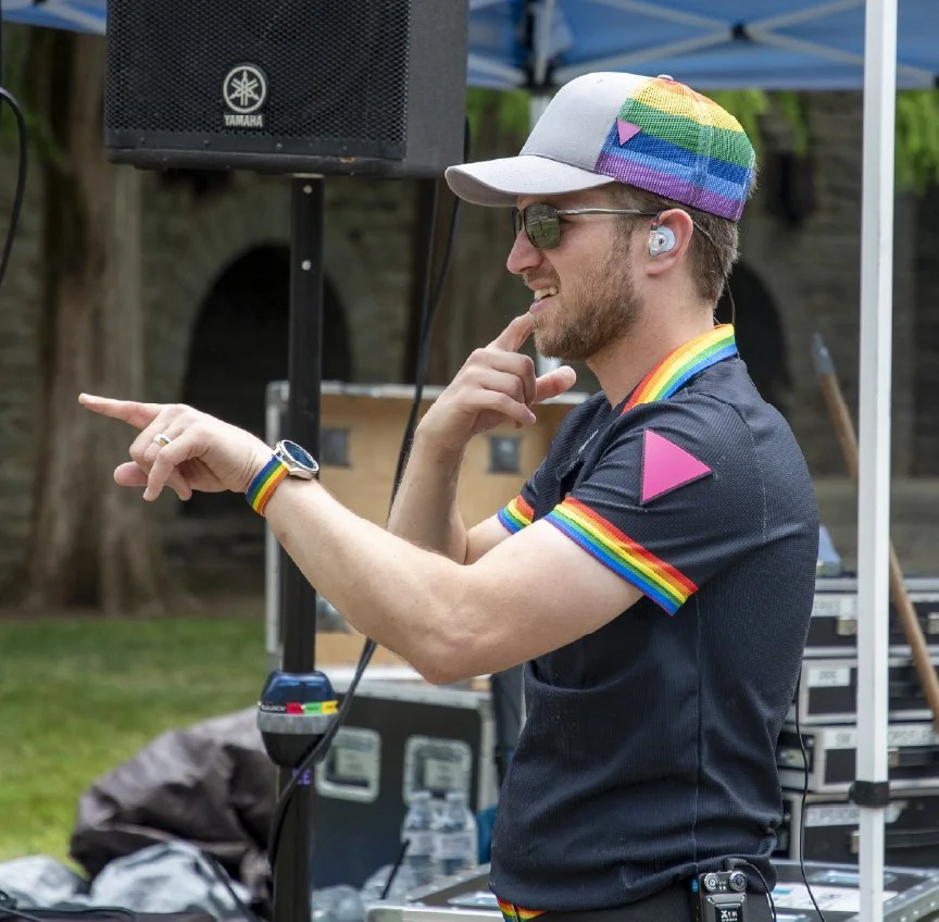 A person at an outdoor event wearing a rainbow pride shirt and accessories, with a rainbow rainbow hat, black sunglasses, and hearing aids. They are smiling and pointing, surrounded by event equipment and trees.