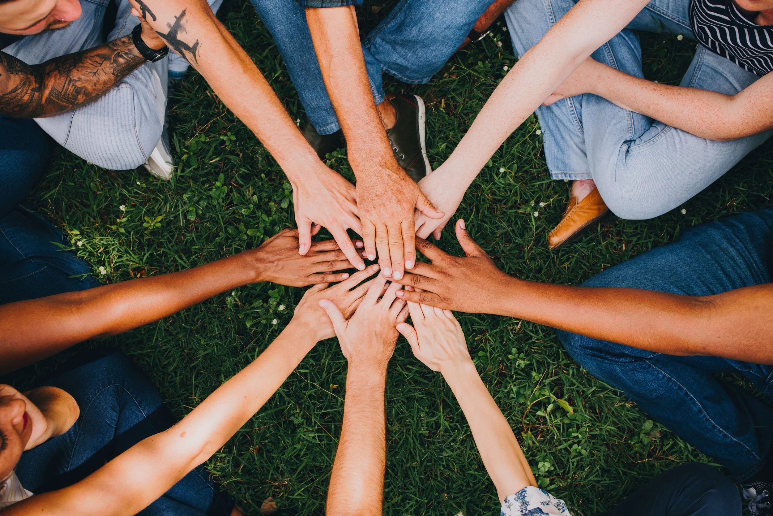 Multiple people with different skin tones forming a circle and stacking their hands in the center, sitting on grass.