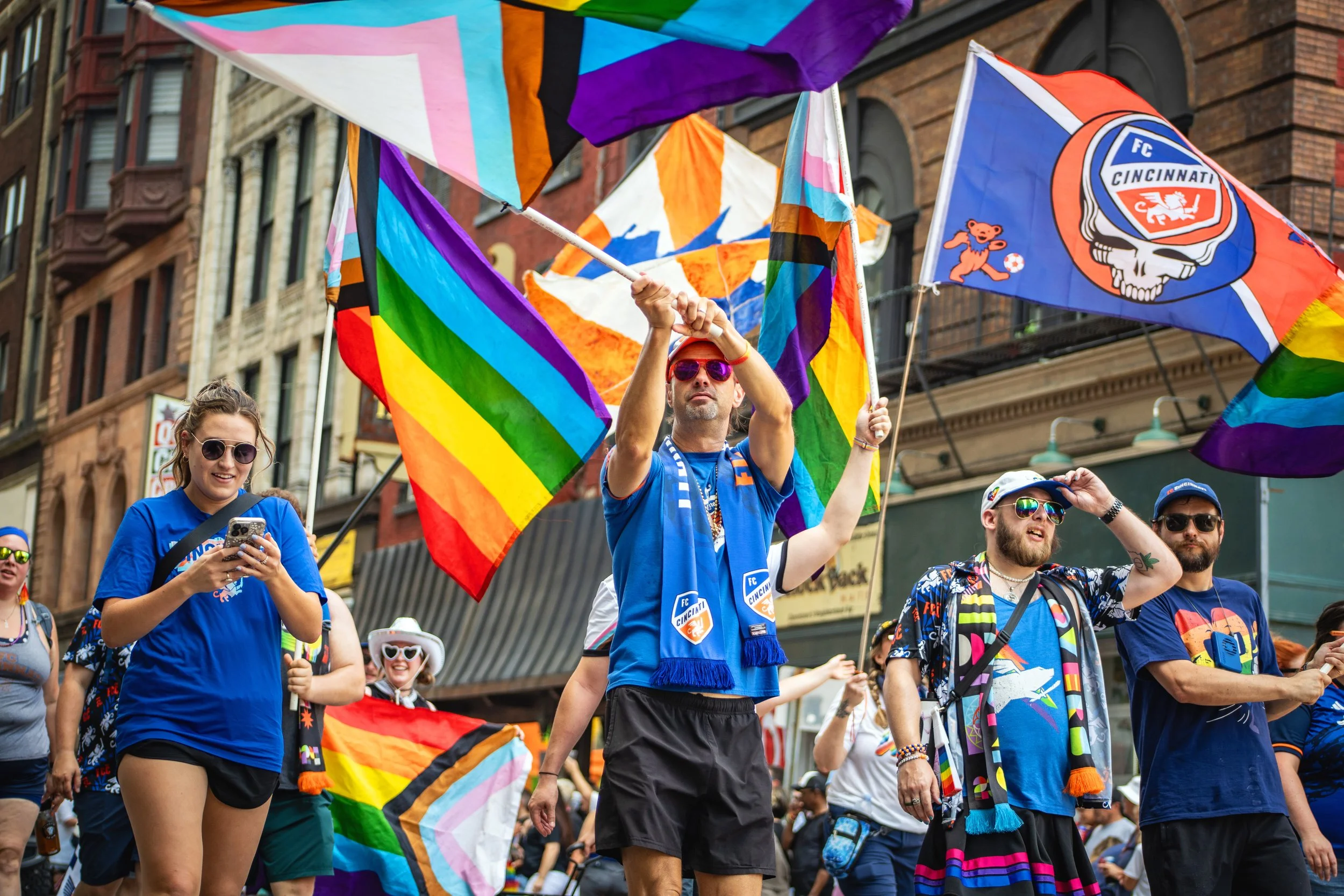 People at a parade holding rainbow pride flags, with some wearing colorful clothes and accessories, in an urban street setting.