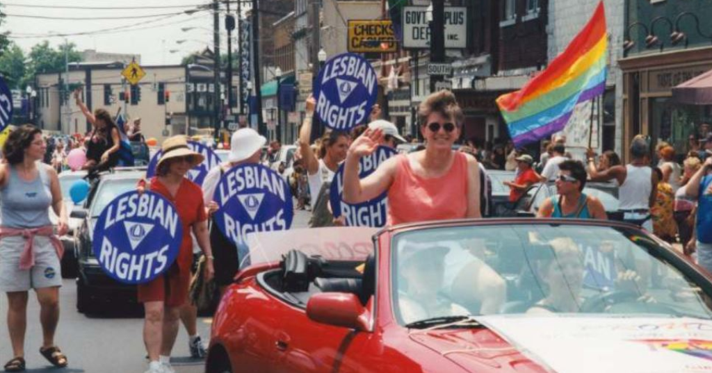 A parade with people holding Lesbian Rights signs and rainbow flags, with some participants on a convertible car and others walking behind on a city street.