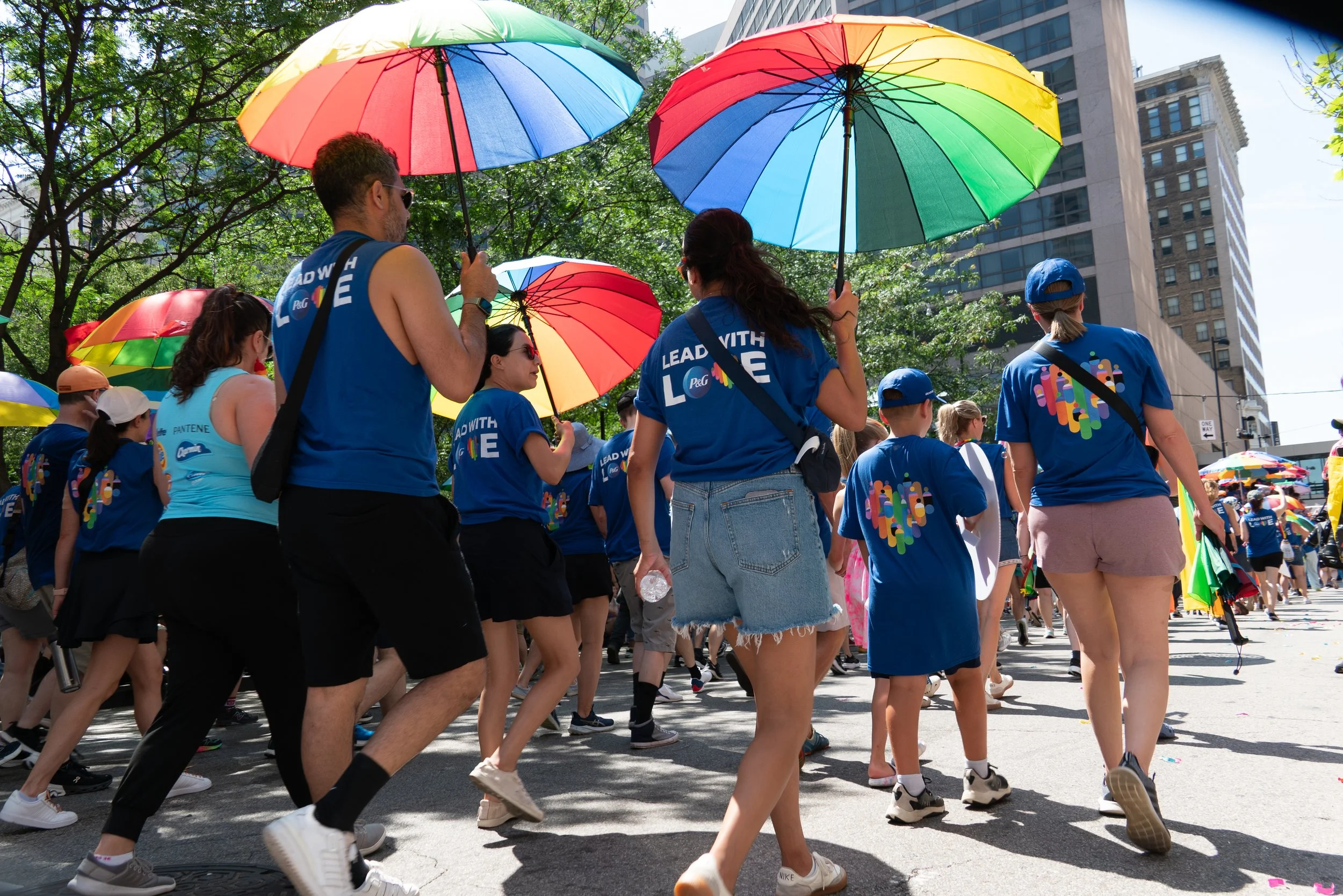 People participating in a parade holding rainbow-colored umbrellas, wearing blue shirts, and walking down a city street with tall buildings and green trees.